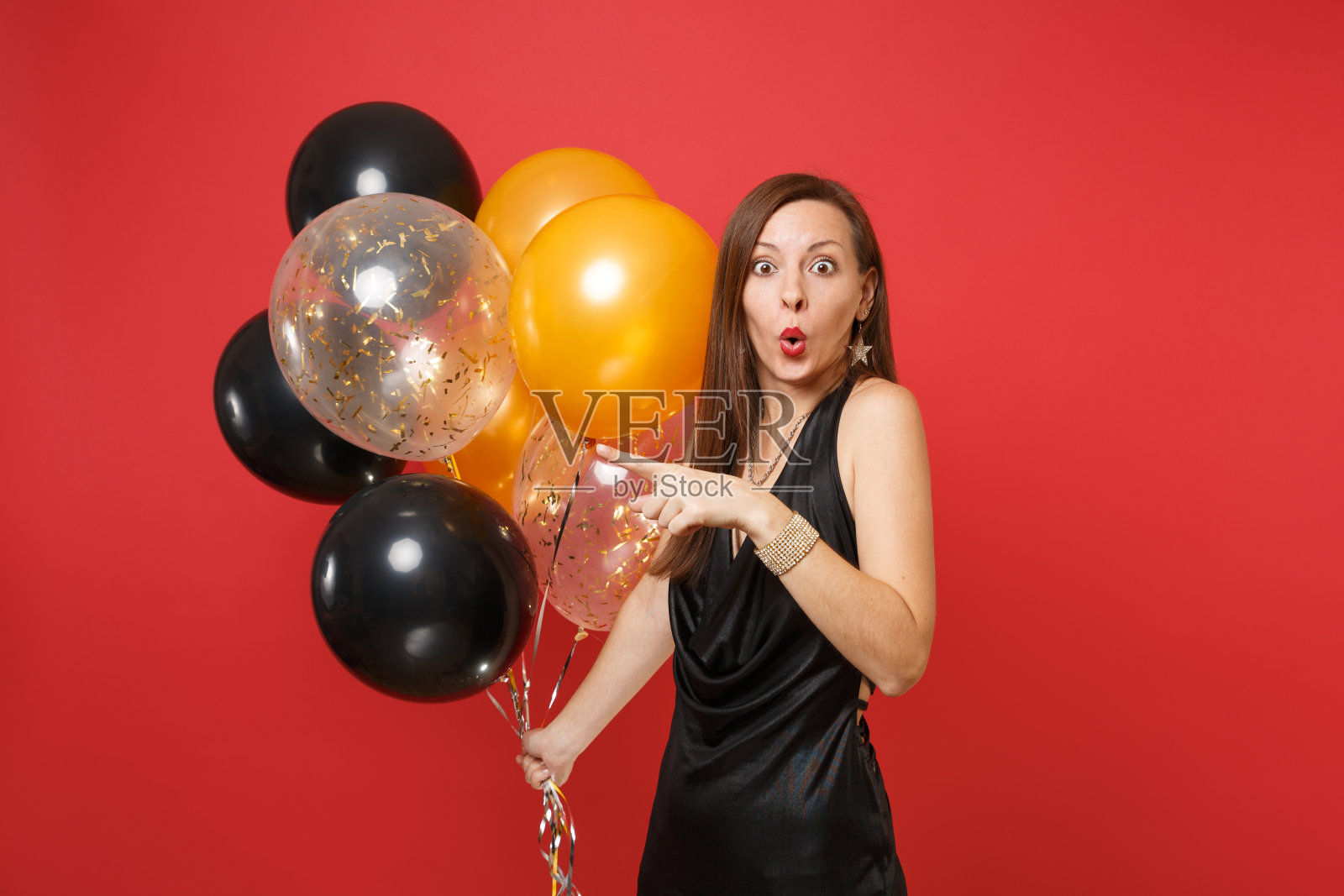 震惊的年轻女子在黑色连衣裙指着食指一边举着气球庆祝孤立的红色背景。国际妇女节新年生日模型节日派对概念。照片摄影图片