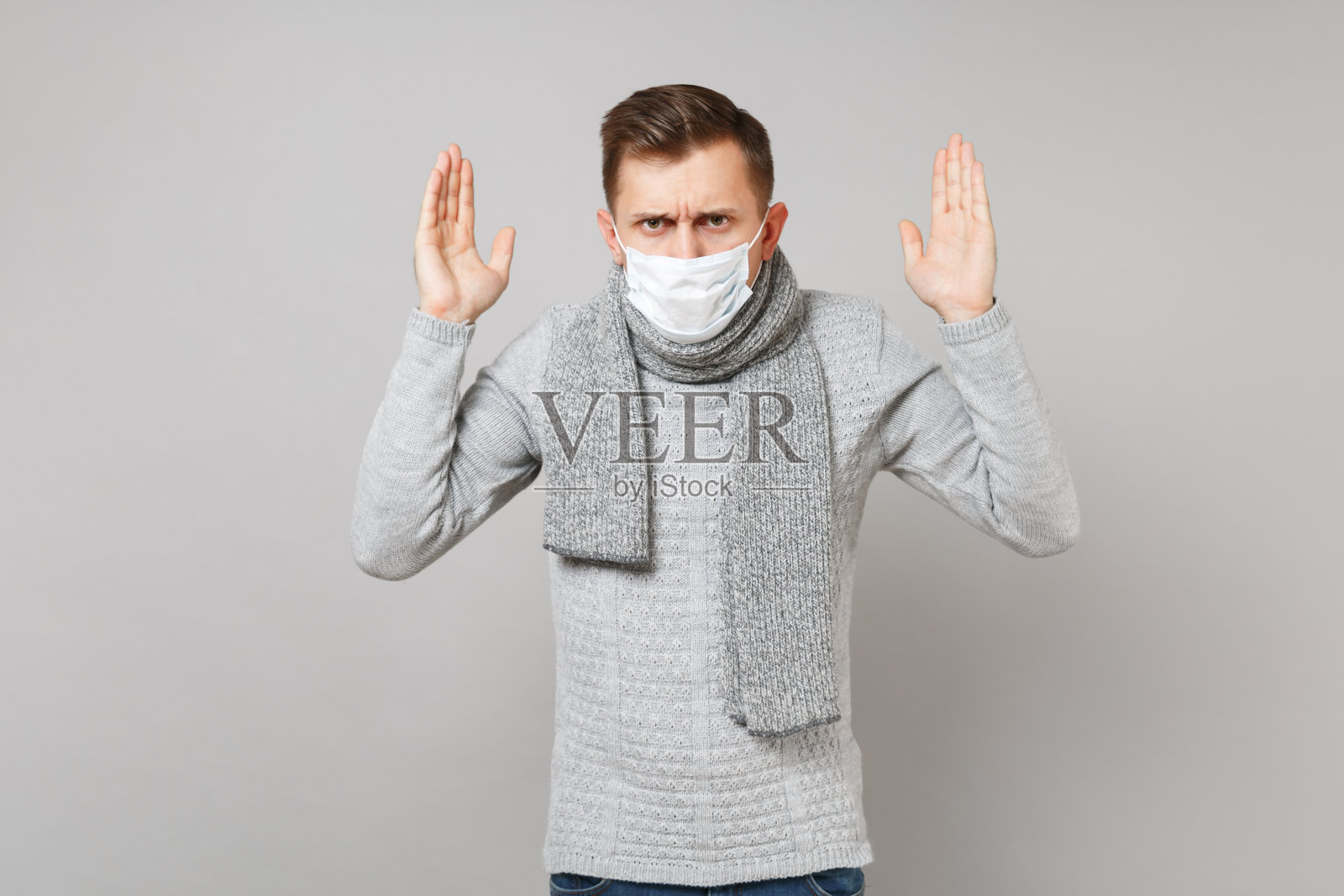 愁眉苦脸的年轻人穿着灰色的毛衣,围着无菌的围巾戴着口罩,双手孤立在灰色的背景上。健康的生活方式,生病生病的疾病治疗,寒冷季节的概念。模拟拷贝空间。照片摄影图片