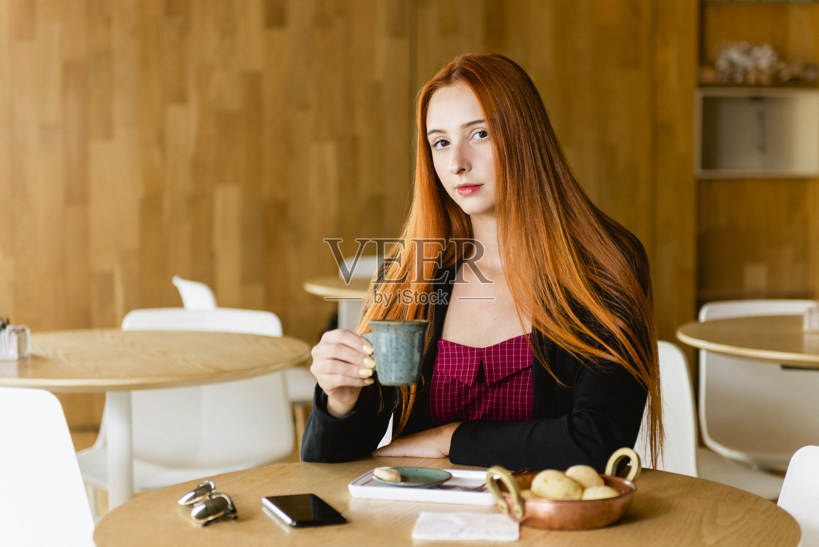 咖啡店里拿着杯子的漂亮红发模特。桌子上放着手机、眼镜和奶酪卷,背景模糊不清照片摄影图片