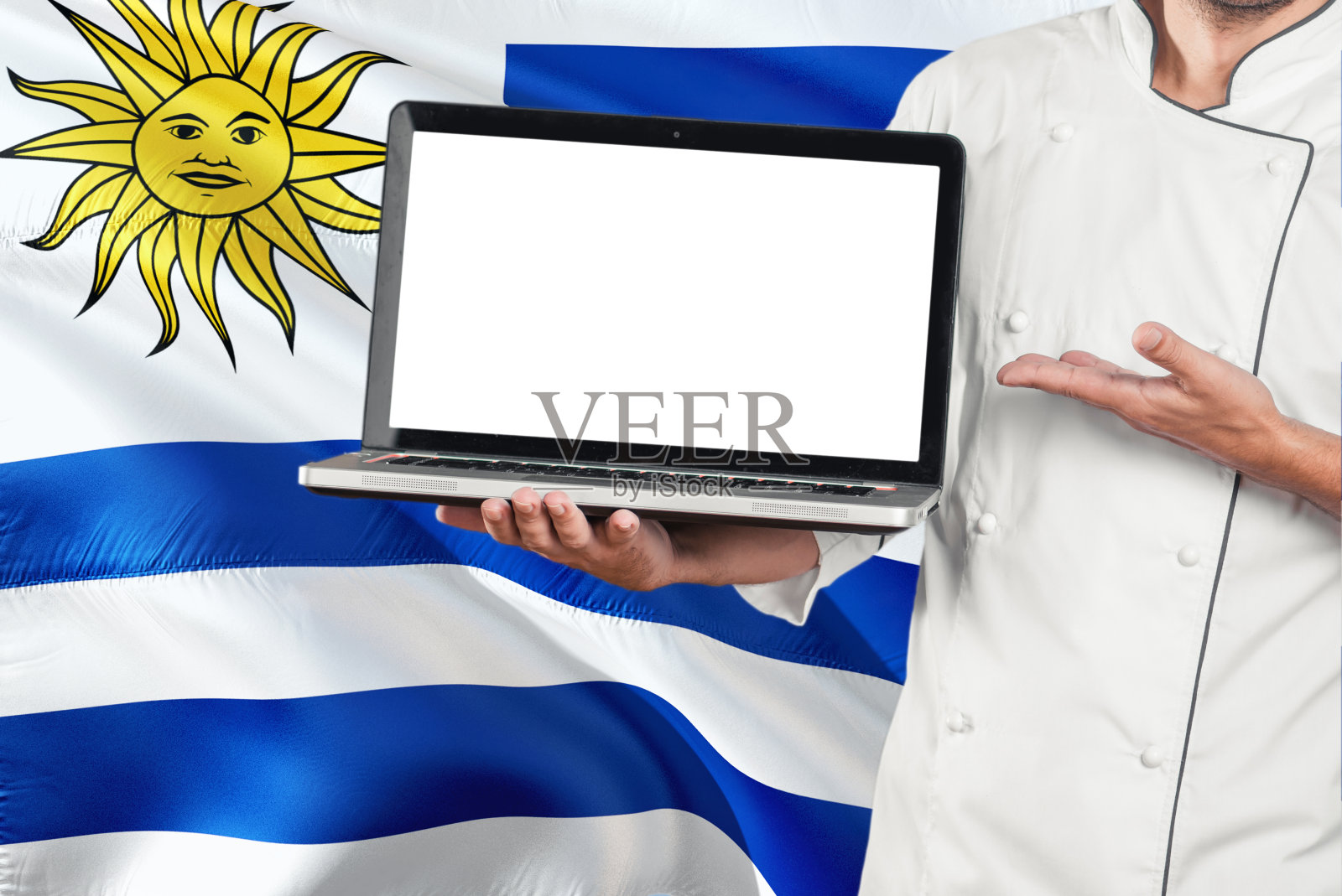 乌拉圭厨师拿着笔记本电脑与空白屏幕上的乌拉圭国旗背景。库克穿着制服,指着笔记本电脑找拷贝空间。照片摄影图片
