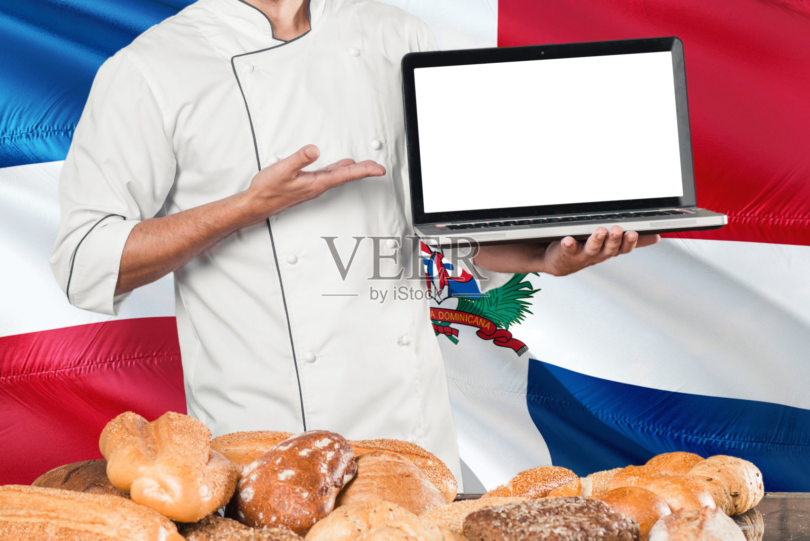 多米尼加面包师拿着笔记本电脑在多米尼加共和国的旗帜和面包的背景。厨师穿着制服指向空白屏幕为拷贝空间。照片摄影图片