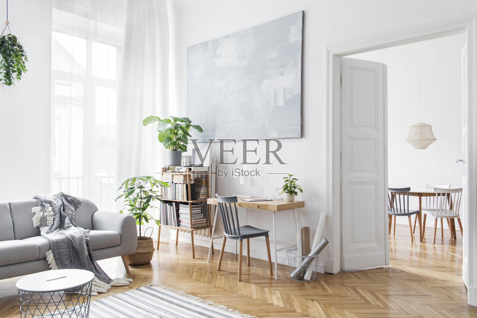 时尚的斯堪的纳维亚开放空间设计家具,植物,竹书架和木制书桌。棕色的木拼花。抽象绘画。现代装饰明亮的房间旁边的餐厅。照片摄影图片