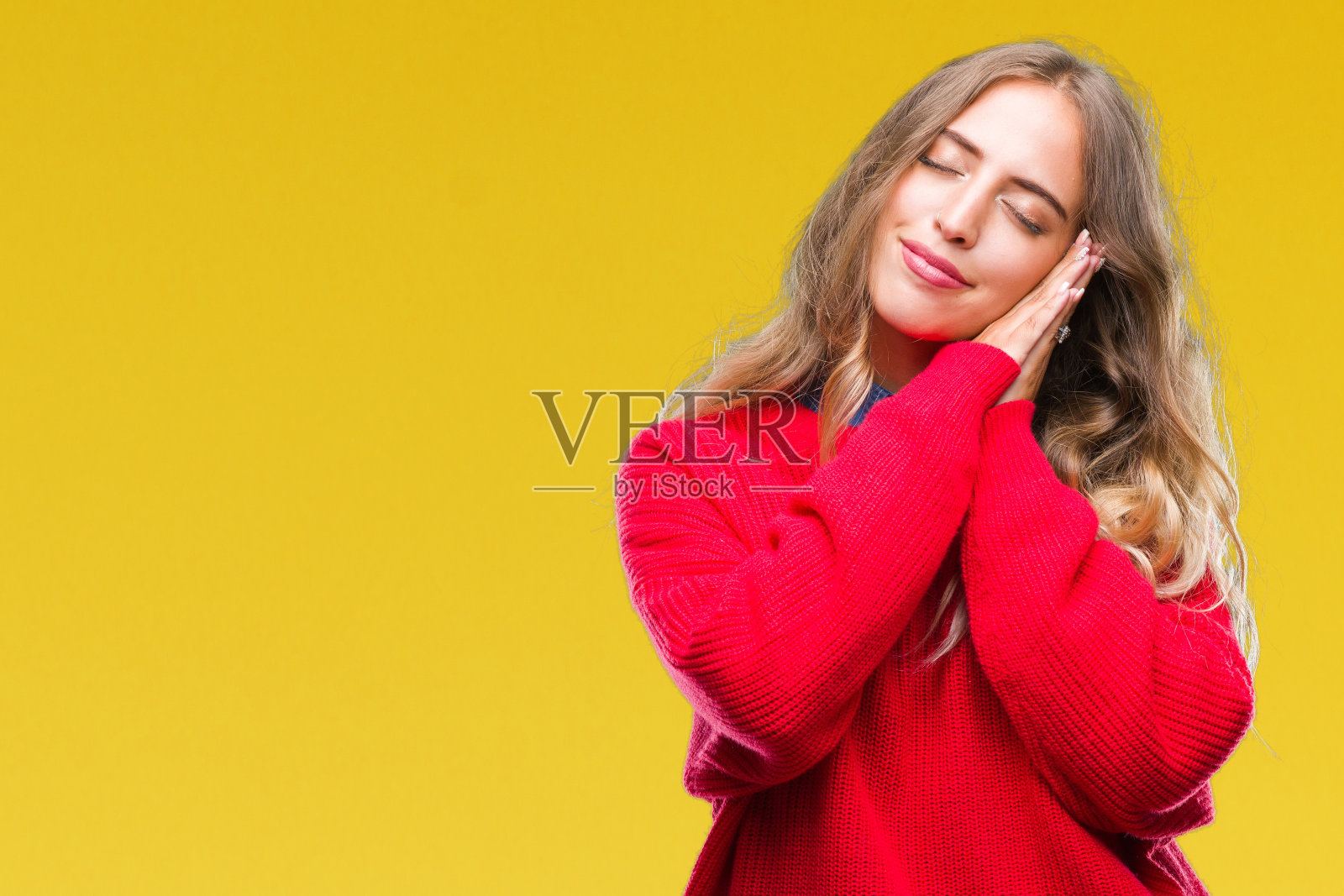 美丽的年轻的金发女人穿着冬天的毛衣在孤立的背景睡累了,做梦,摆姿势与手在一起,而闭上眼睛微笑。照片摄影图片