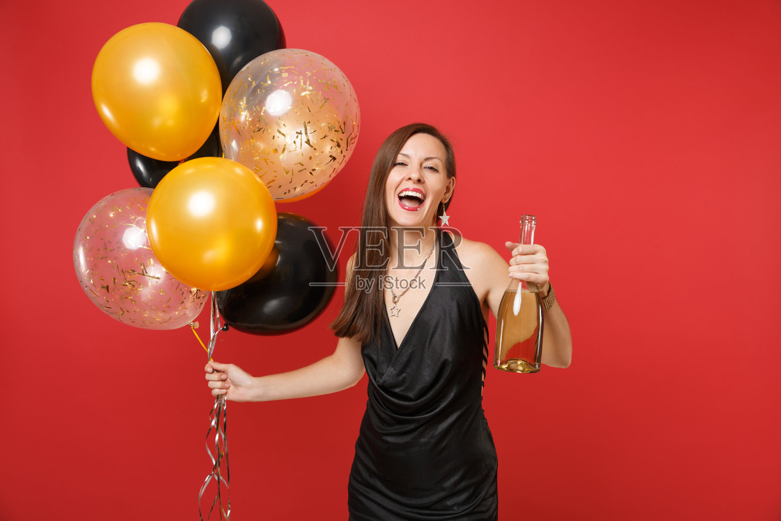 快乐的年轻女孩穿着小黑裙庆祝,拿着一瓶香槟,气球孤立在红色的背景。情人节,新年快乐,生日模型节日派对概念。照片摄影图片