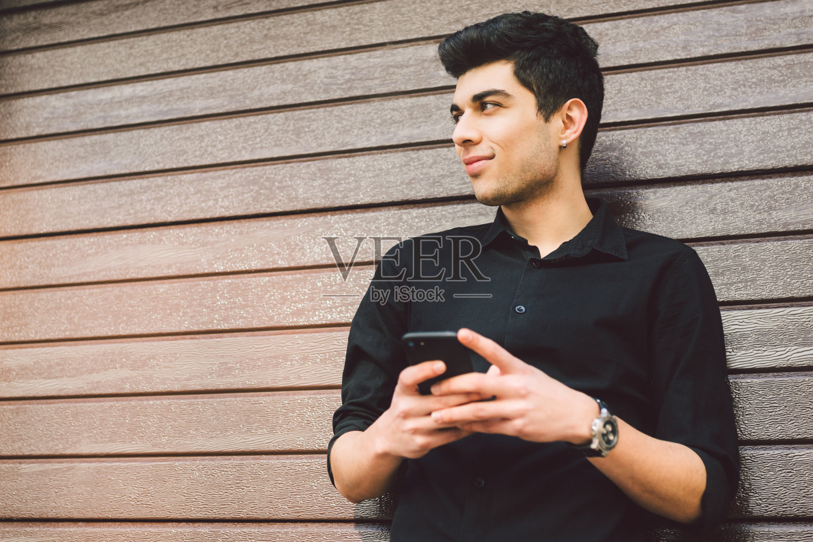 年轻英俊的土耳其男模特肖像。在阳光明媚的天气里,一名身穿黑色衬衫的地中海裔黑发女子正在街道上的木墙背景上使用一名科技商人的手机照片摄影图片