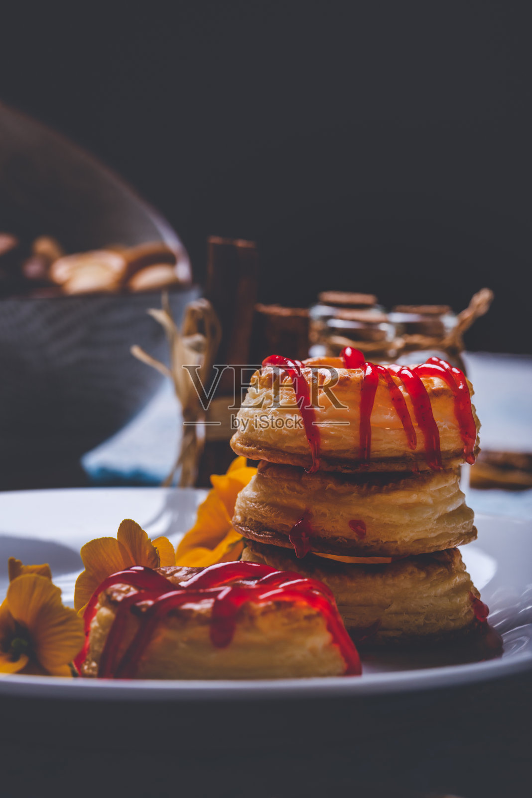 用油酥面团做成的几个甜甜圈的垂直照片。红色草莓酱洒在戒指上。甜甜圈放在白色的盘子上,有少量黄色的花。照片摄影图片