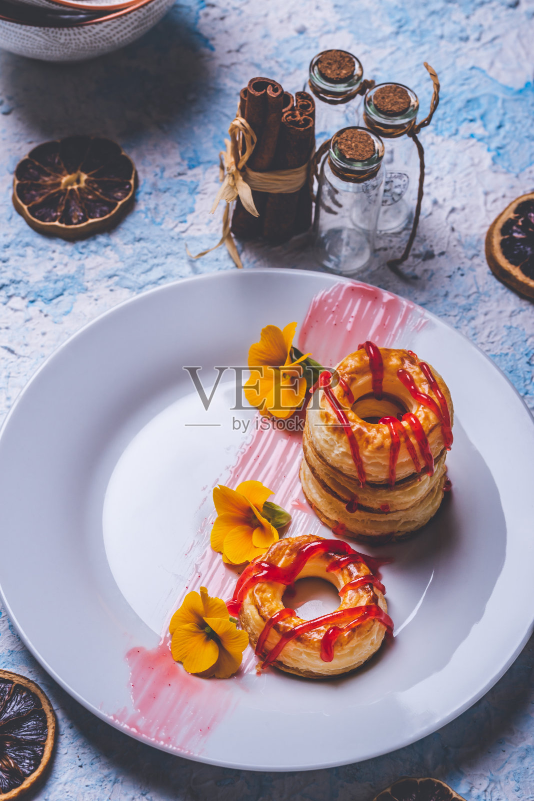 用油酥面团做成的几个甜甜圈的垂直照片。红色草莓酱洒在戒指上。甜甜圈放在白色的盘子上,有少量黄色的花。照片摄影图片