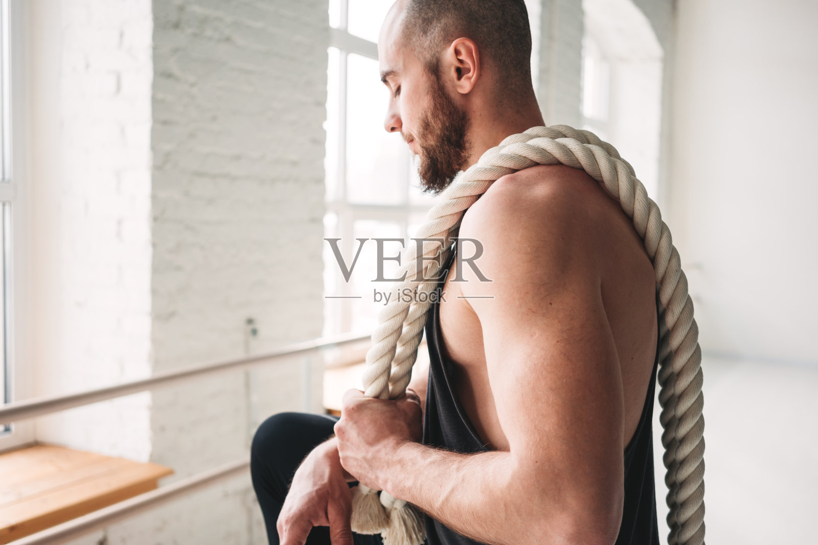 英俊的肌肉男子抱着肩绳在轻健身房锻炼。健身男子的肖像与战斗绳在交叉健身房照片摄影图片