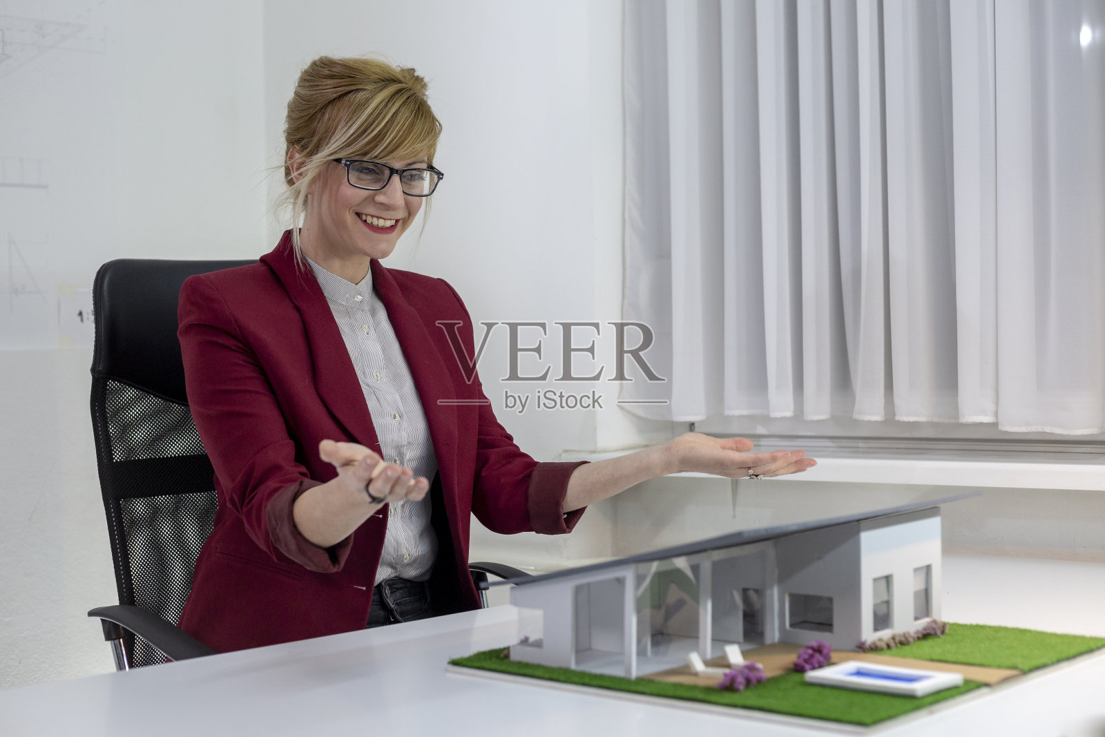 年轻女建筑师在办公室坐在桌子上看房子模型。深思熟虑的设计师戴着眼镜思考项目。照片摄影图片