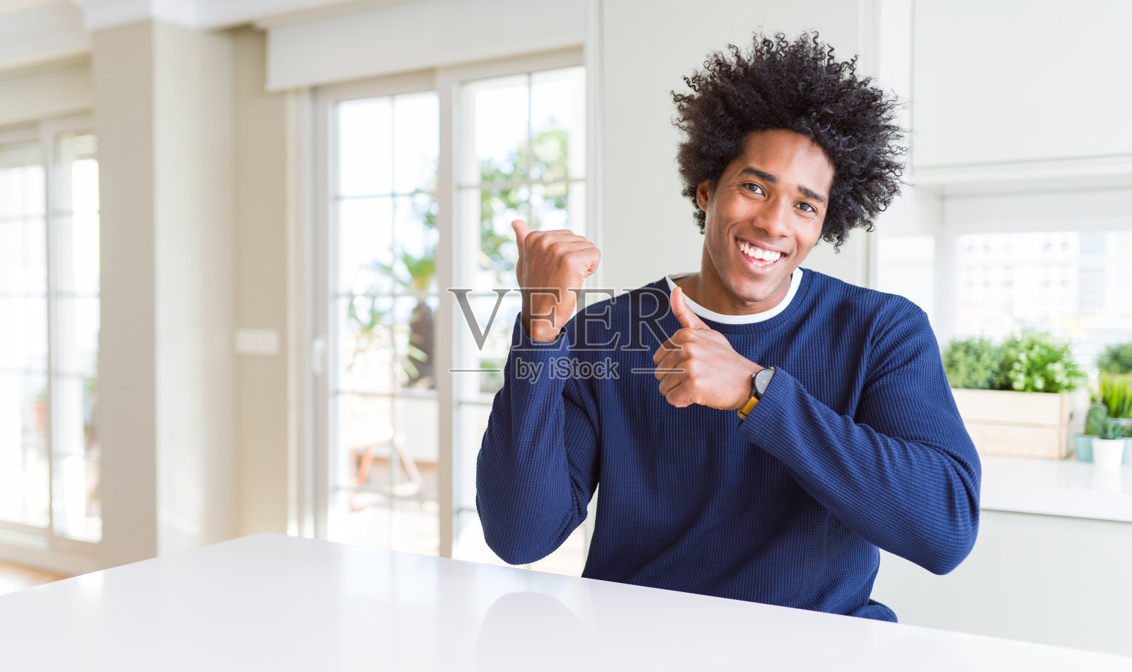 年轻的非洲裔美国人穿着休闲毛衣坐在家里,用手和拇指指着背后,自信地微笑照片摄影图片