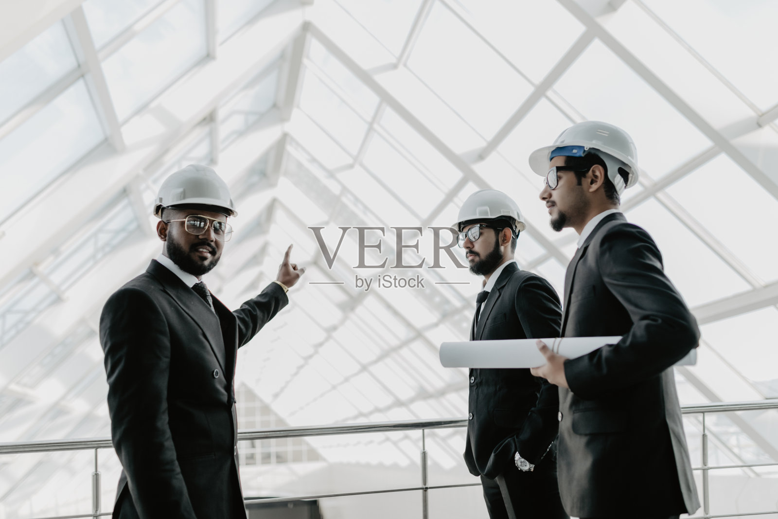 年轻严肃的印度男子戴着头盔,打着领带,带着平板电脑,陪同两名工程师指着建筑管道。管制员检查新大楼。会议与建设理念照片摄影图片