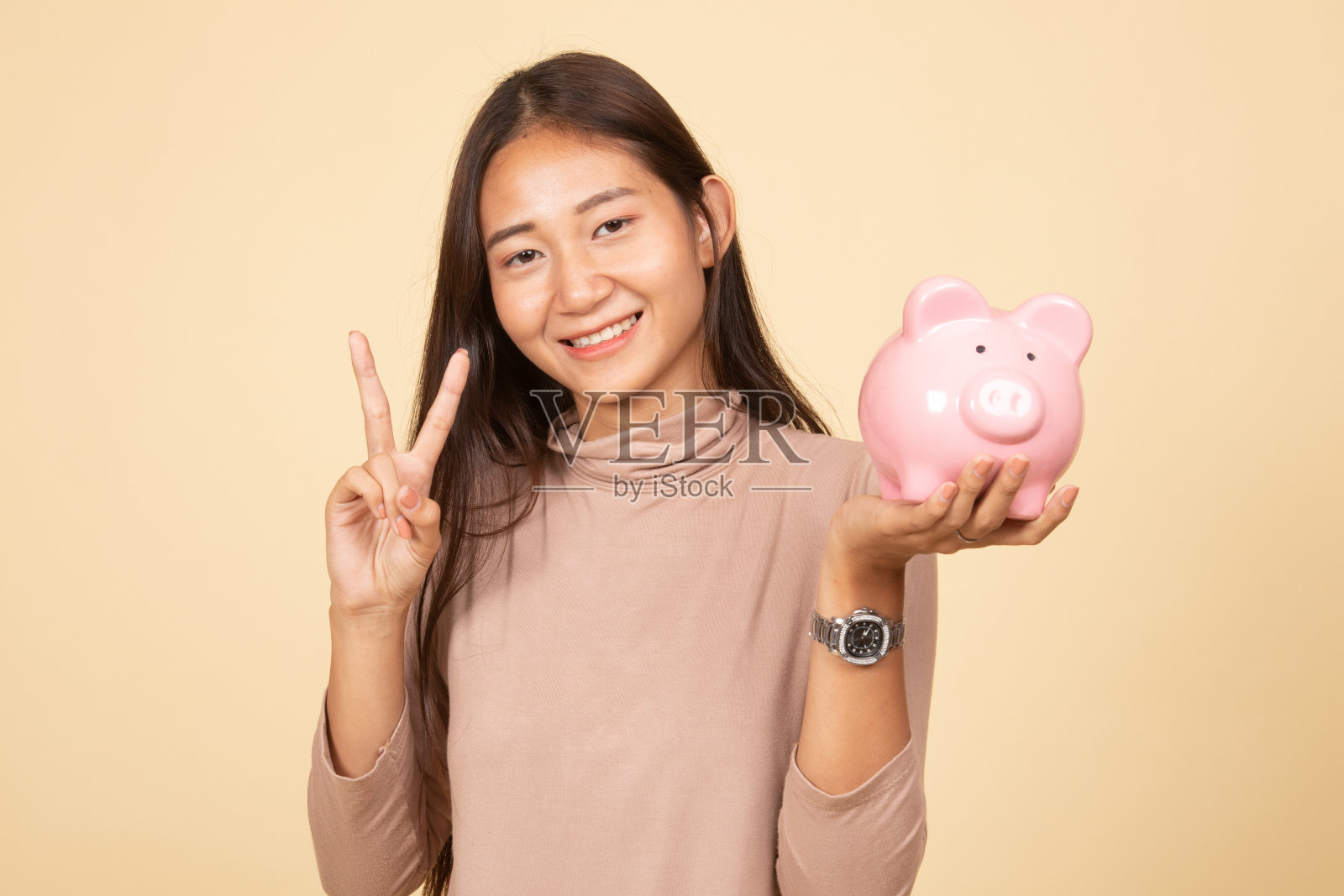 亚洲妇女用猪硬币银行显示胜利的标志。照片摄影图片