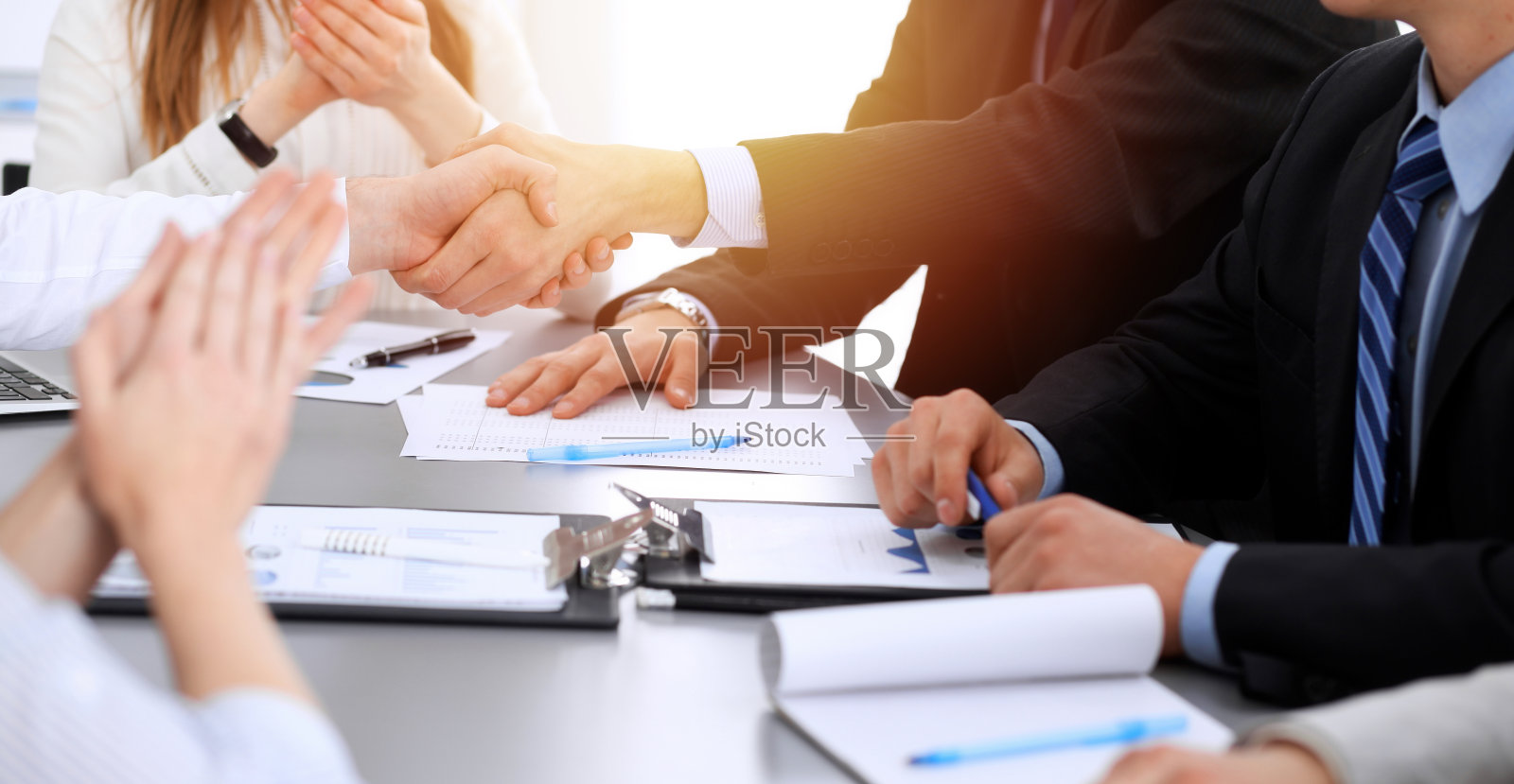 商务人士在会议上握手,而他们的同事在鼓掌和鼓掌。一群不知名的商人和妇女在现代白人办公室。成功的团队合作、伙伴关系和握手理念照片摄影图片