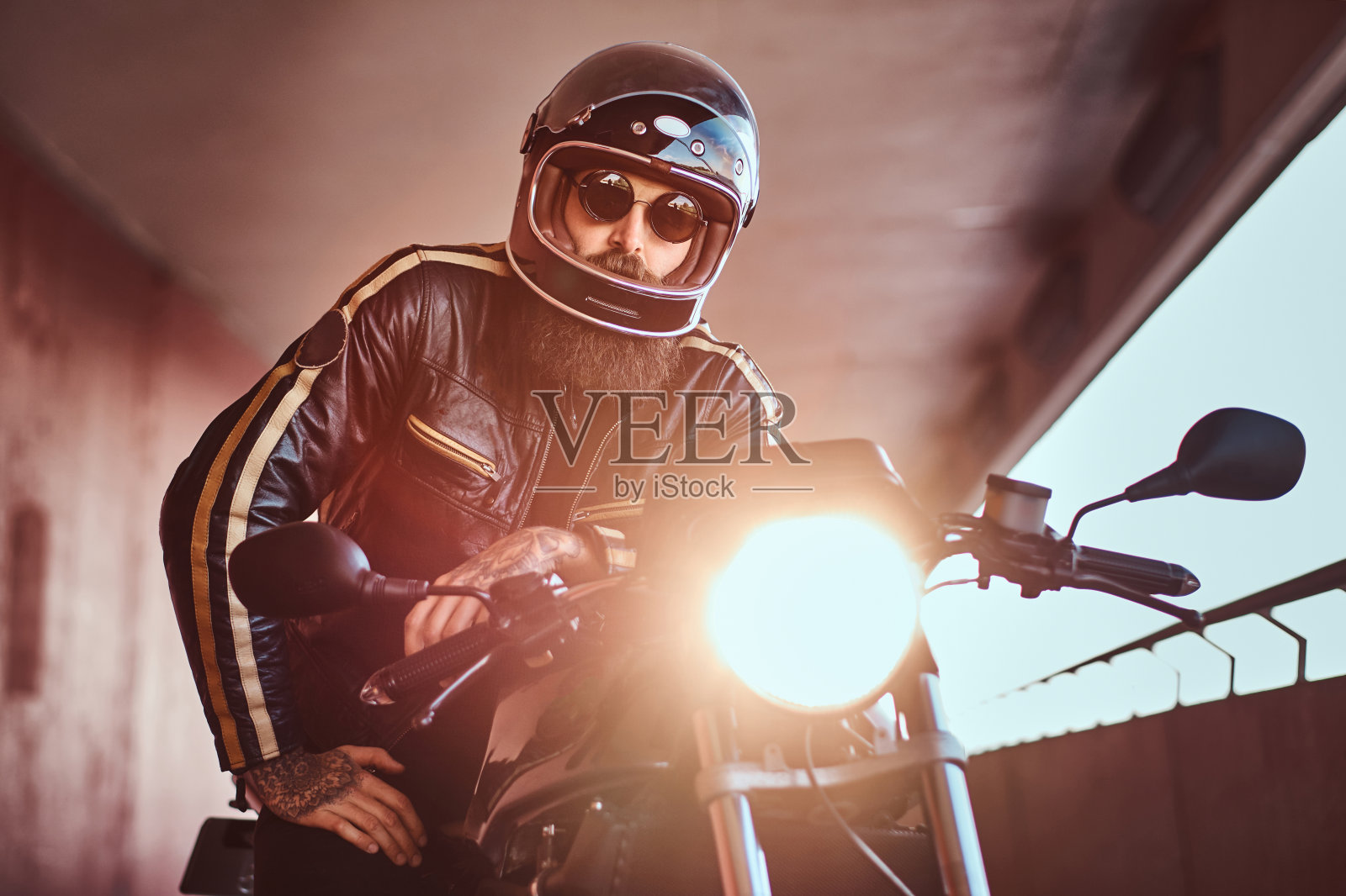 近距离的肖像,一个野蛮的大胡子摩托车手戴着头盔和太阳镜,穿着黑色皮夹克坐在一个复古摩托车与包括前灯。照片摄影图片