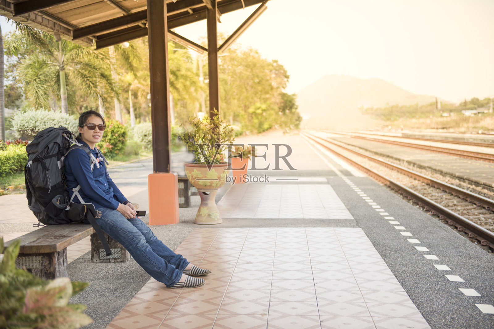 快乐的亚洲背包女子在休闲服装坐在火车站和手持智能手机。照片摄影图片