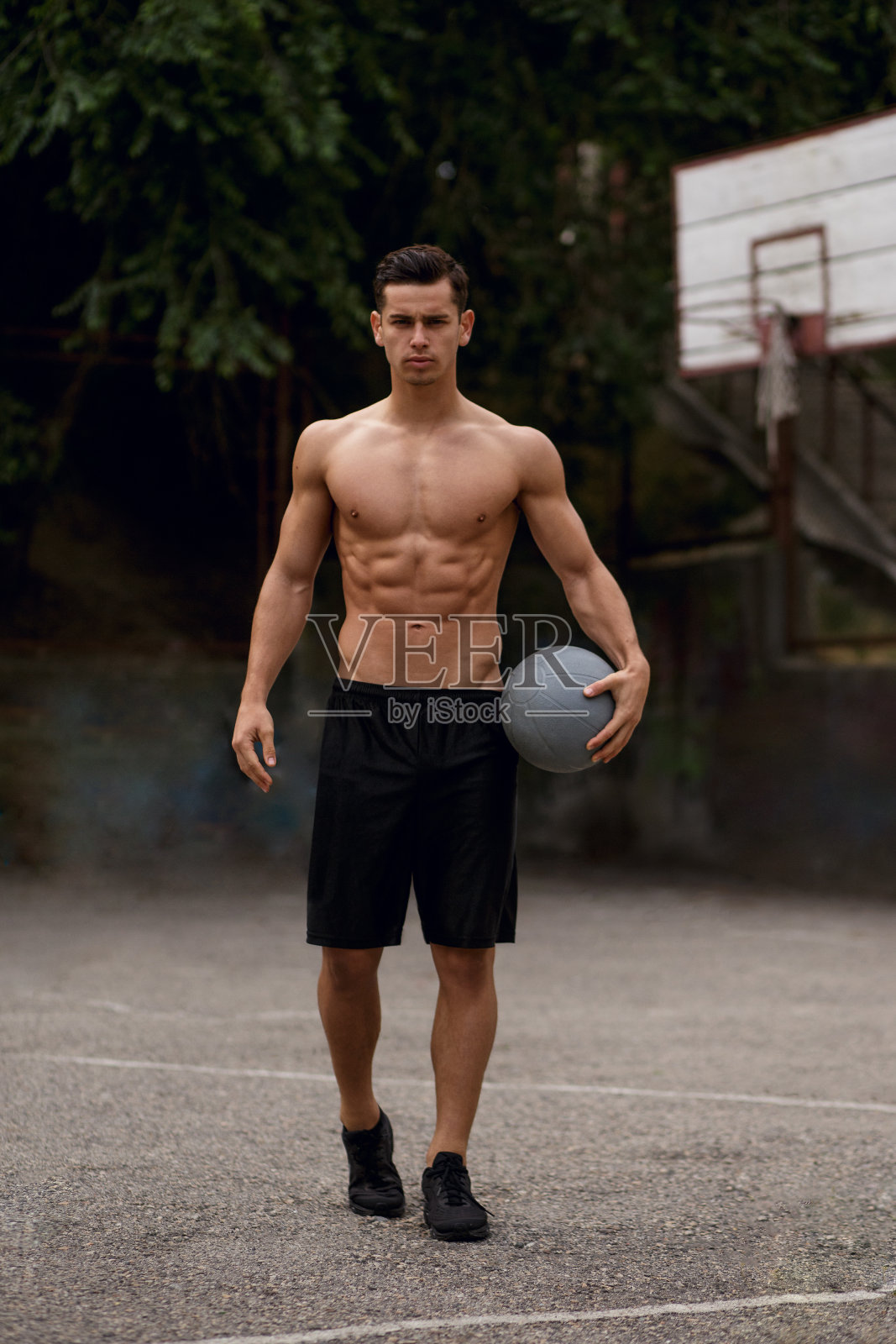 赤裸上身,运动健将,肌肉发达,手里拿着球,坐在篮球场上,孤立地站在街道上。照片摄影图片