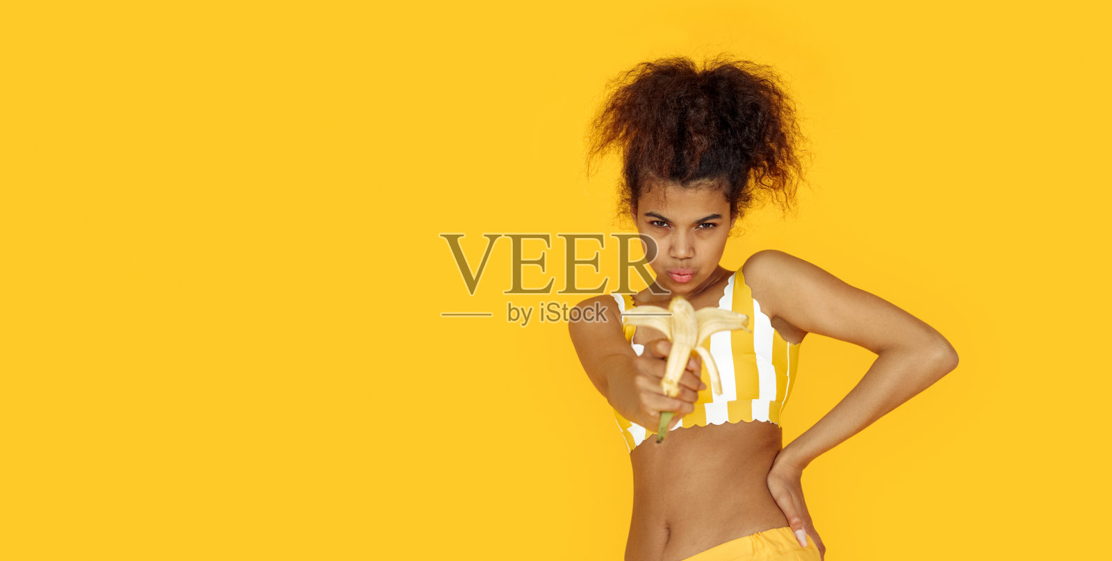 自信年轻的非洲裔美国女人假装拿着香蕉枪看着相机,酷的黑人少女使射击姿态孤立在夏季黄色工作室背景,肖像,横幅设计照片摄影图片