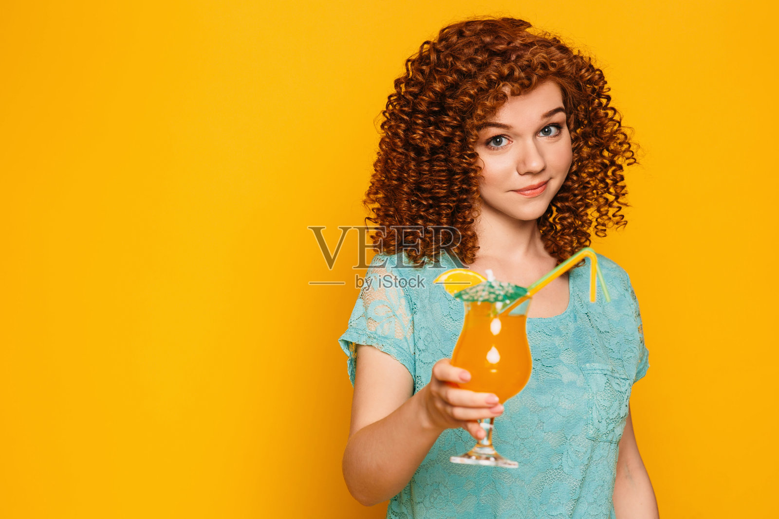 一头红发的卷发女人,黄色背景上是一杯夏日鸡尾酒。照片摄影图片