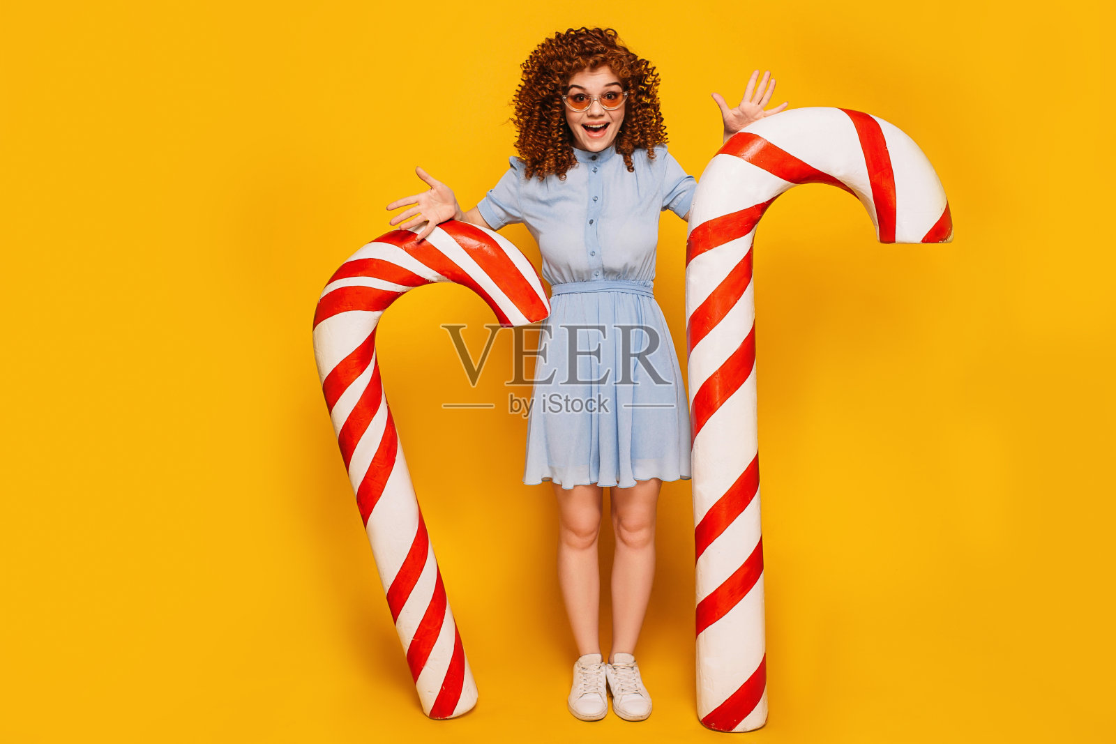 卷曲的红头发积极的女人站在甜美的大糖果拐杖在黄色的背景照片摄影图片