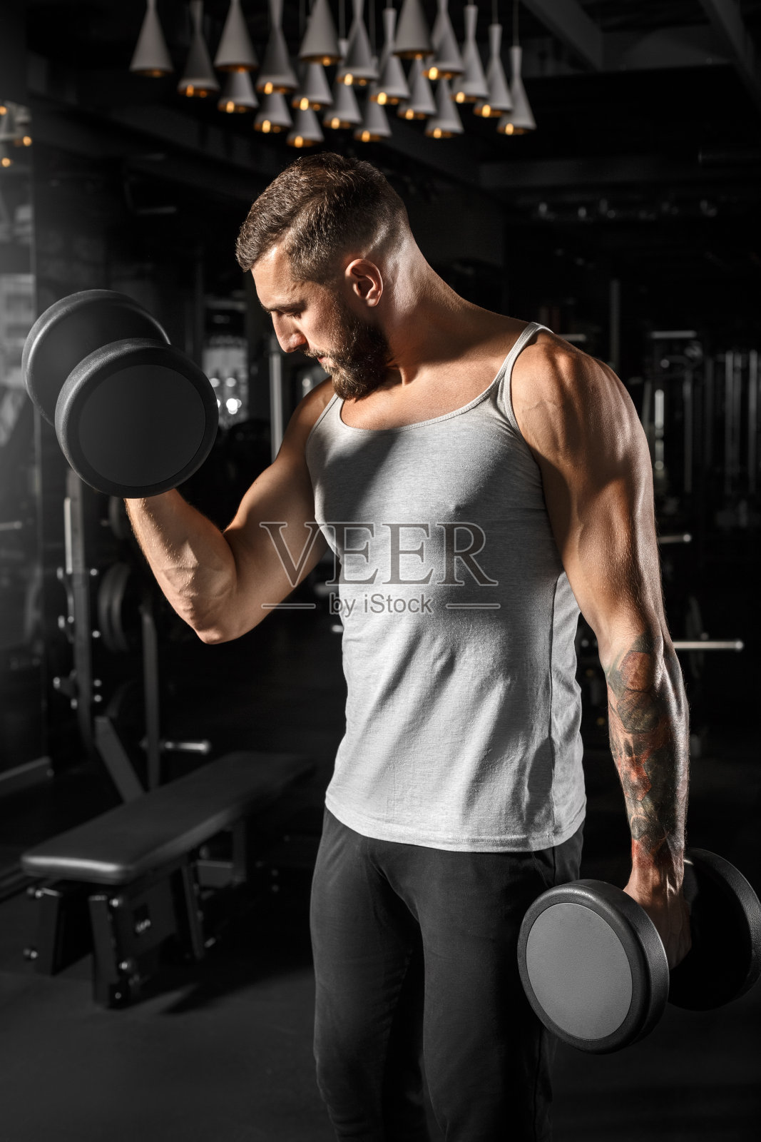 健身。一个留着胡子的男人拿着哑铃站在健身房,好奇地看着二头肌照片摄影图片