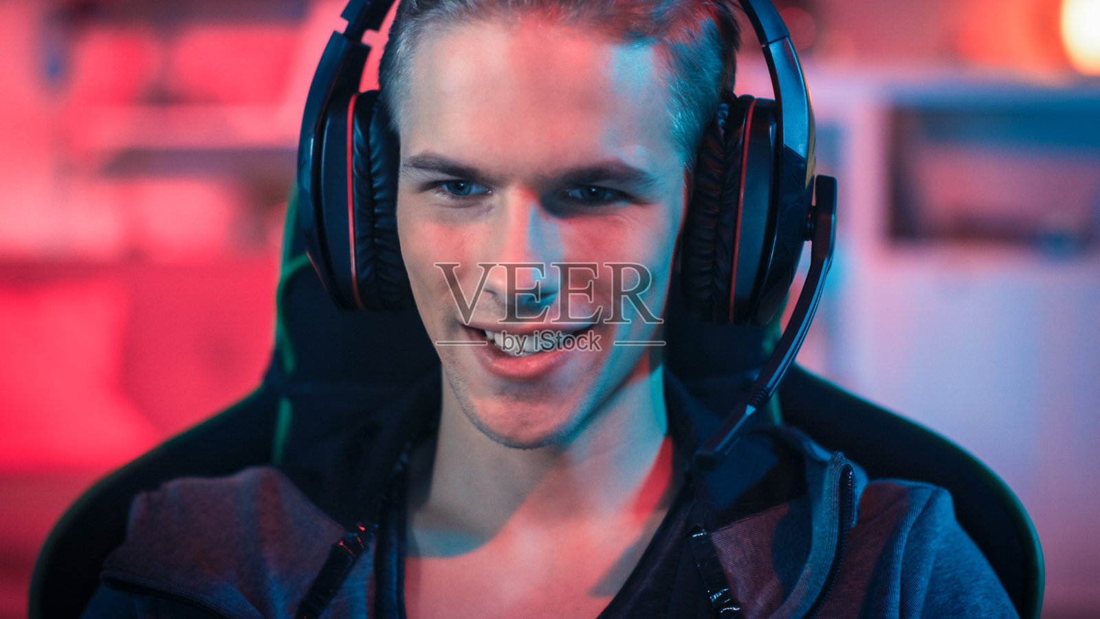 戴着麦克风在个人电脑上玩在线视频游戏的玩家。他邪恶地微笑。房间和个人电脑有红色霓虹灯Led灯。温馨的家庭之夜。照片摄影图片