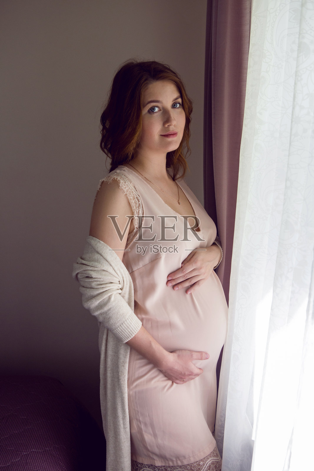 穿着粉色丝绸睡衣的孕妇女孩站在家里的窗帘大窗前照片摄影图片