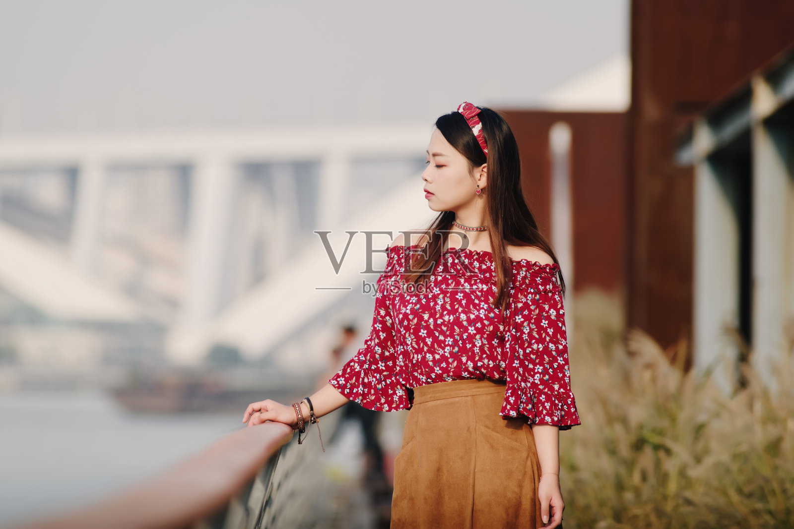 美丽的中国女孩的肖像在一个阳光明媚的秋天的下午,穿着红色的衣服摆姿势在河带与手在栏杆上。照片摄影图片