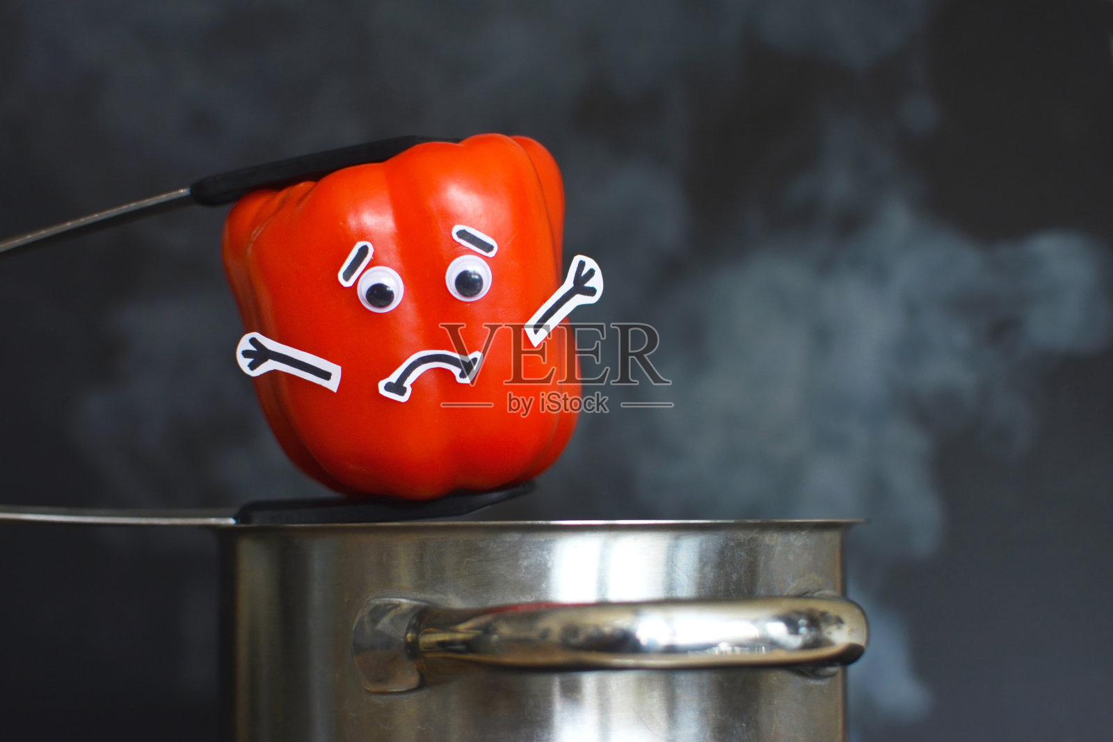 带着悲伤表情和瞪着眼睛的红辣椒被放入一个冒着热气的锅中,锅的背景是黑色的照片摄影图片