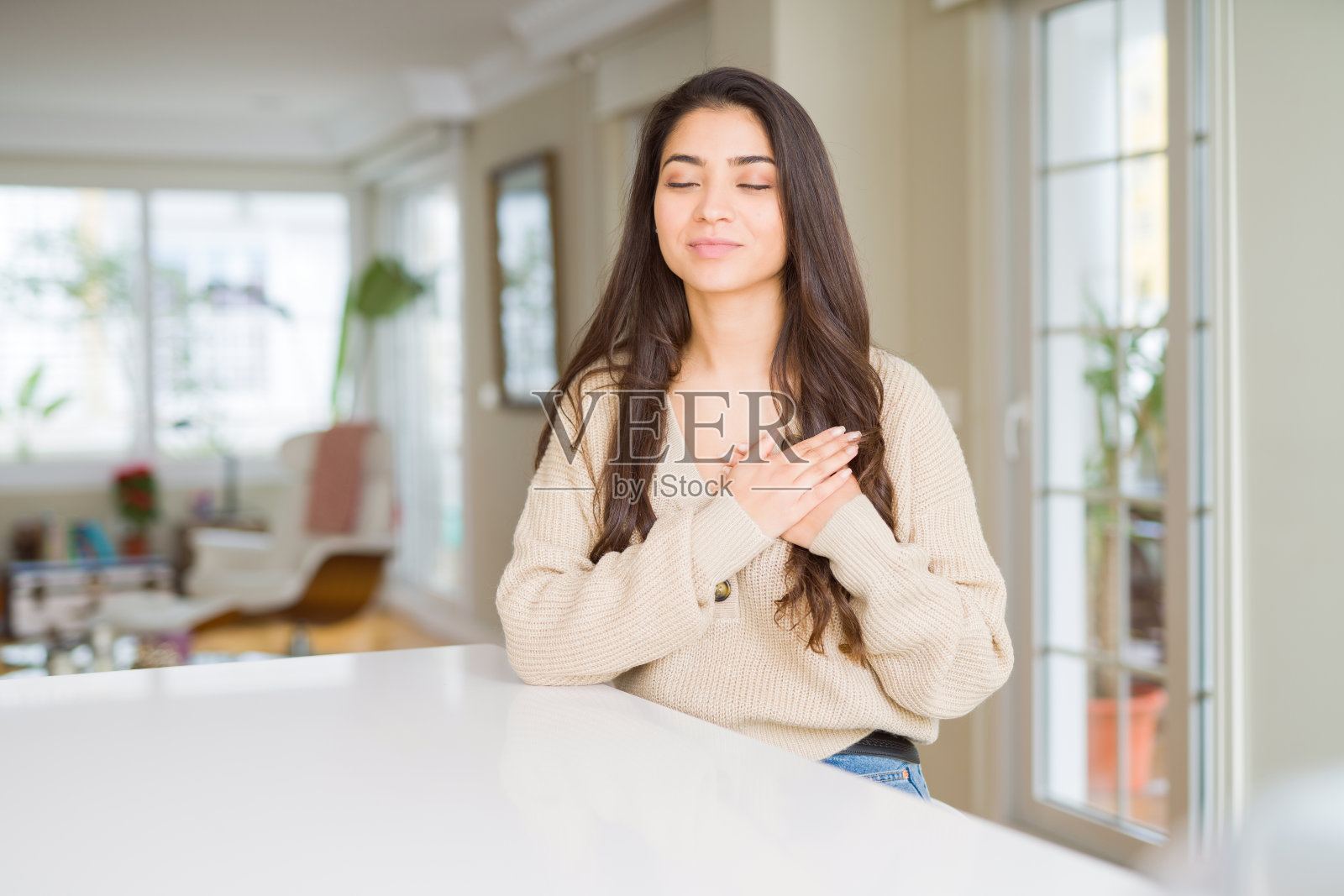 年轻漂亮的女人在家里微笑,双手放在胸前,闭上眼睛,脸上有感激的手势。健康的概念。照片摄影图片