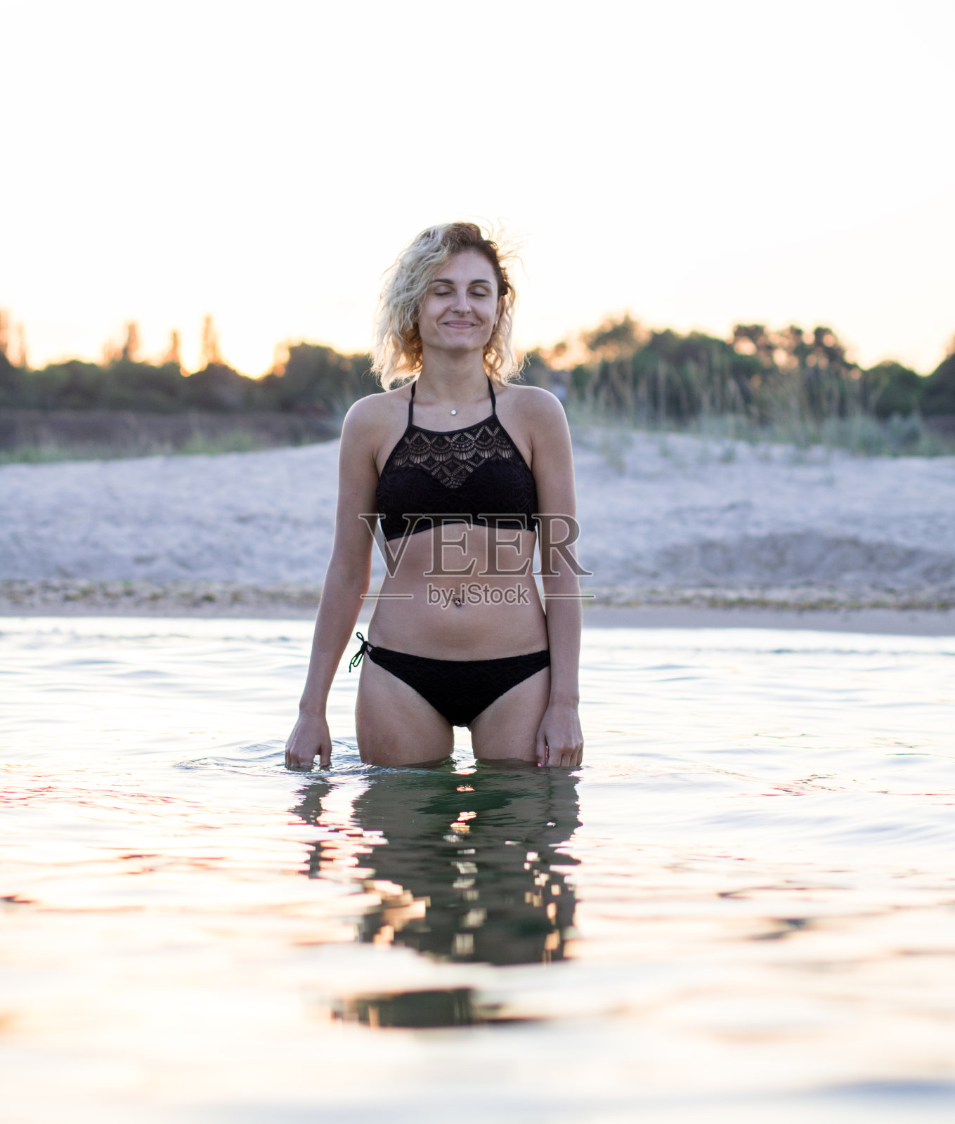 穿着黑色泳衣的卷发女郎正站在沙滩上的水里,夕阳在背景上照片摄影图片