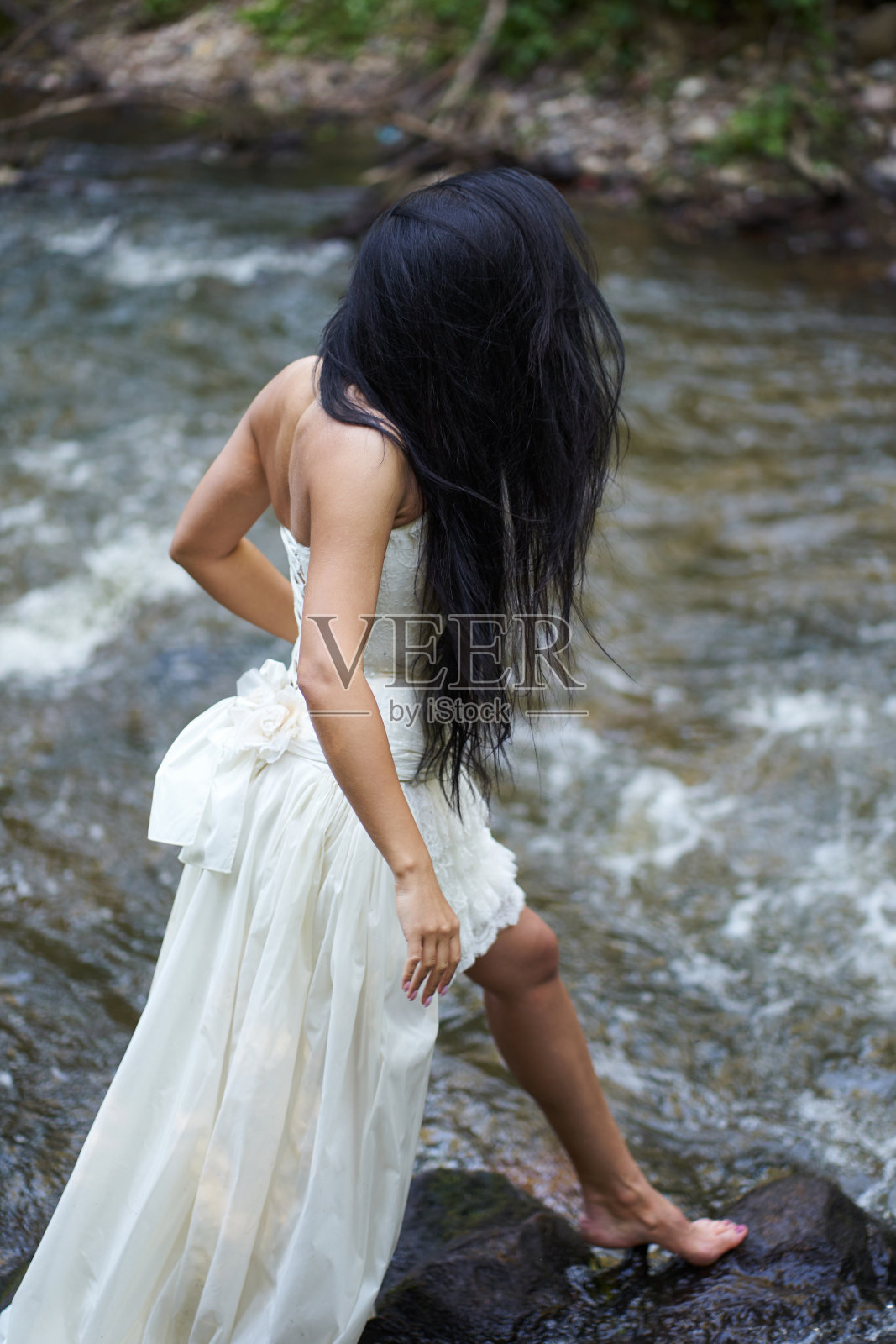 年轻漂亮的黑发女人穿着白色婚纱试图从岩石上过河照片摄影图片