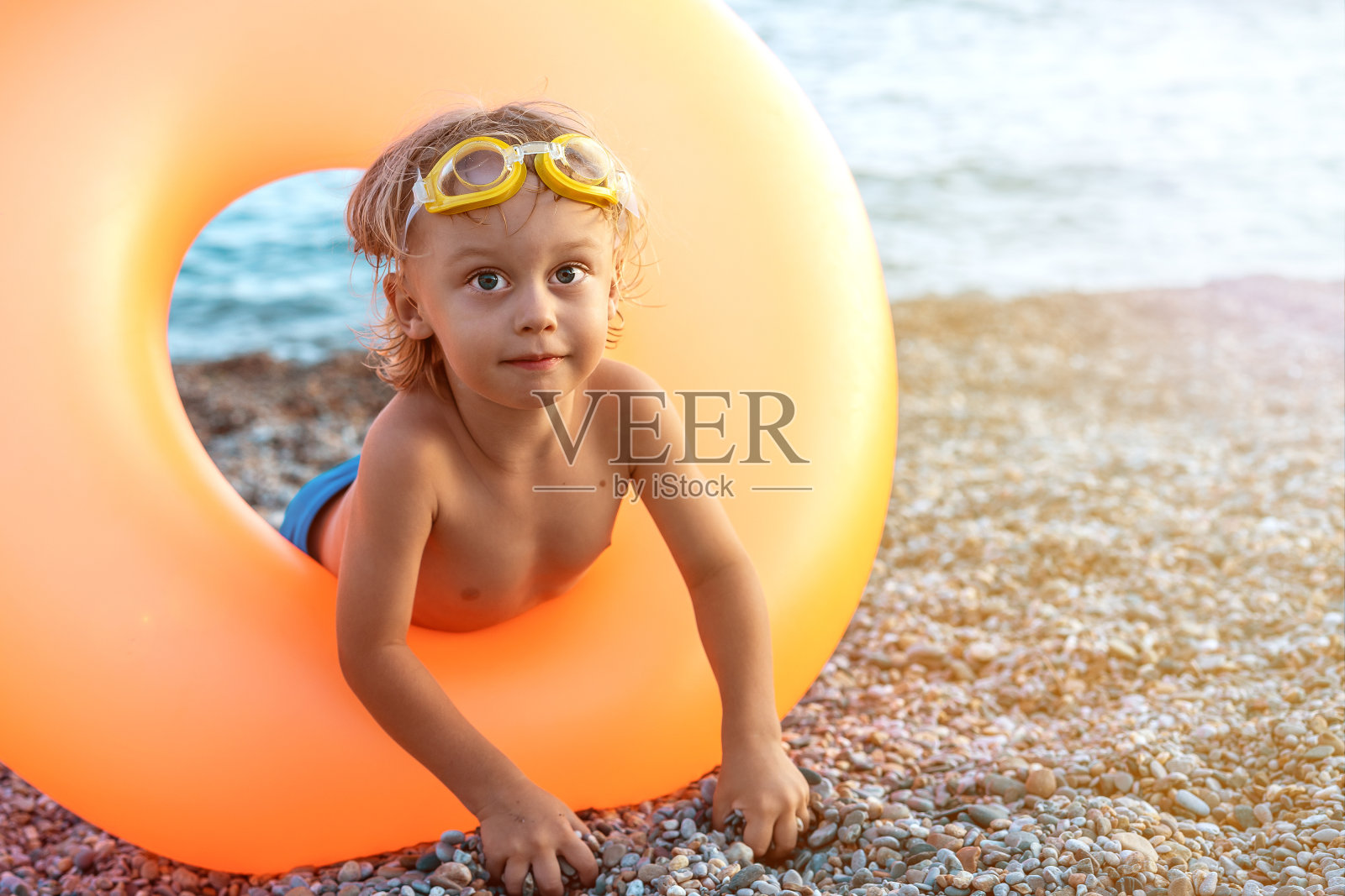漂亮的金发小男孩坐在橡胶轮里在海边玩鹅卵石。假期的概念照片摄影图片