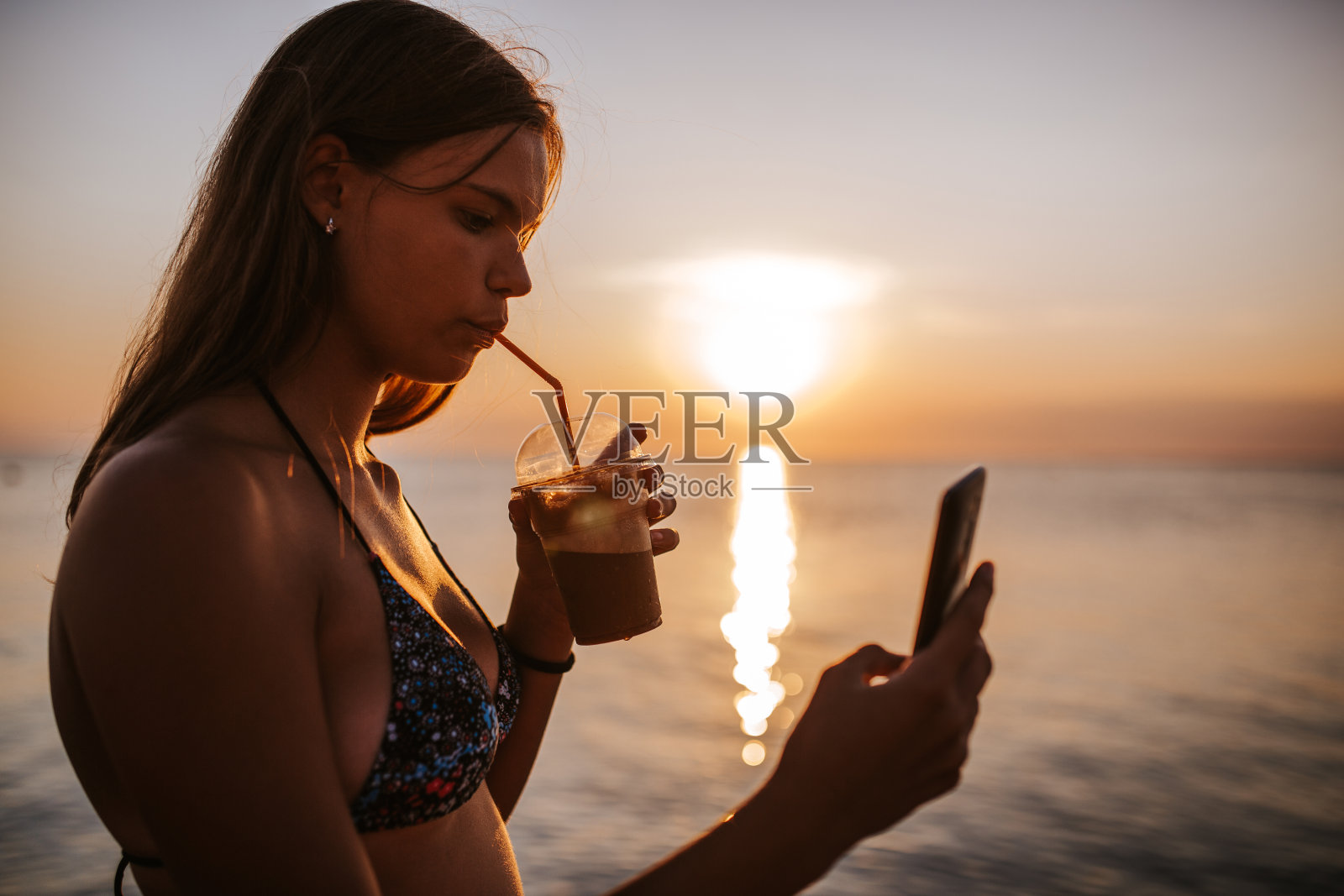 在美丽的夕阳下,十几岁的女孩在海滩上喝着咖啡,用着手机照片摄影图片