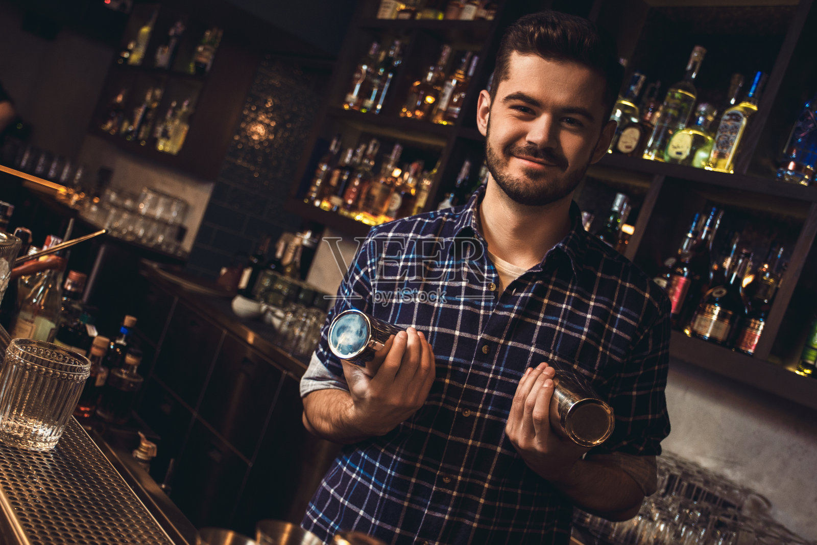 年轻的酒保拿着调酒器,站在吧台前开玩笑照片摄影图片