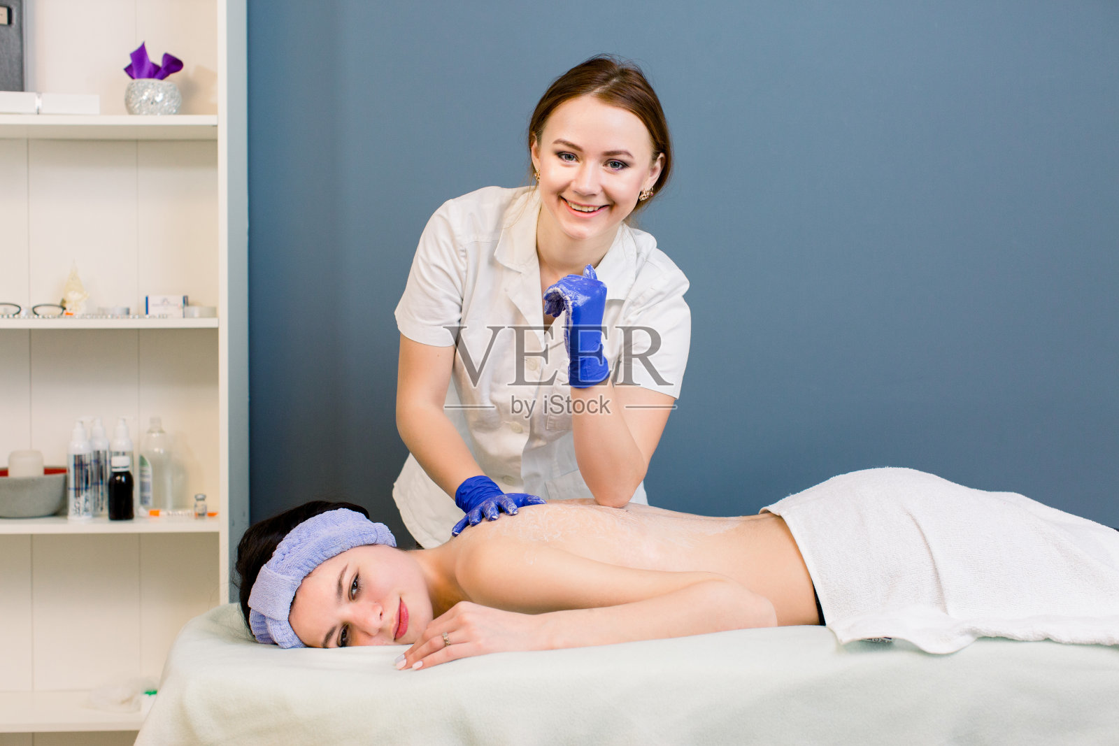 女人拿回水疗沙龙按摩与年轻的美容师。年轻的微笑医学美容师看着相机在工作。照片摄影图片