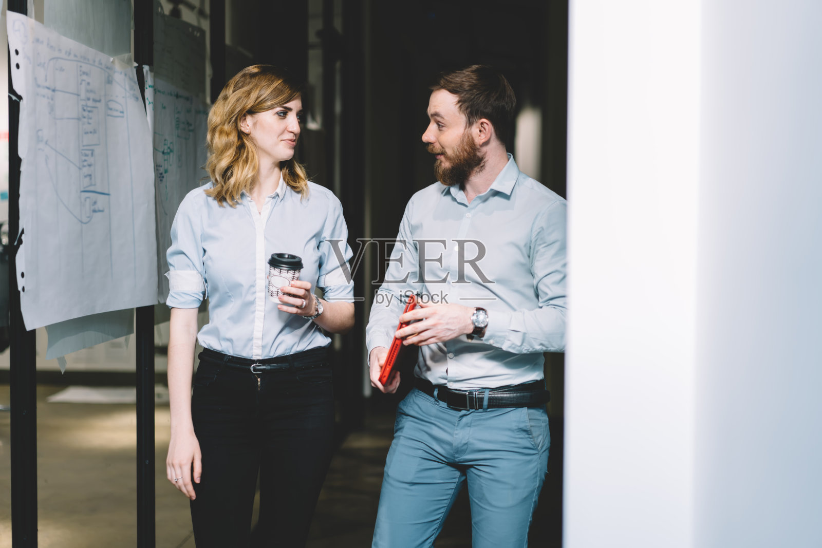 一个神奇的年轻人手里拿着笔记本电脑,和拿着咖啡走在公共区域附近的走廊上的女同事聊天。惊讶的男员工讨论工作时间表照片摄影图片