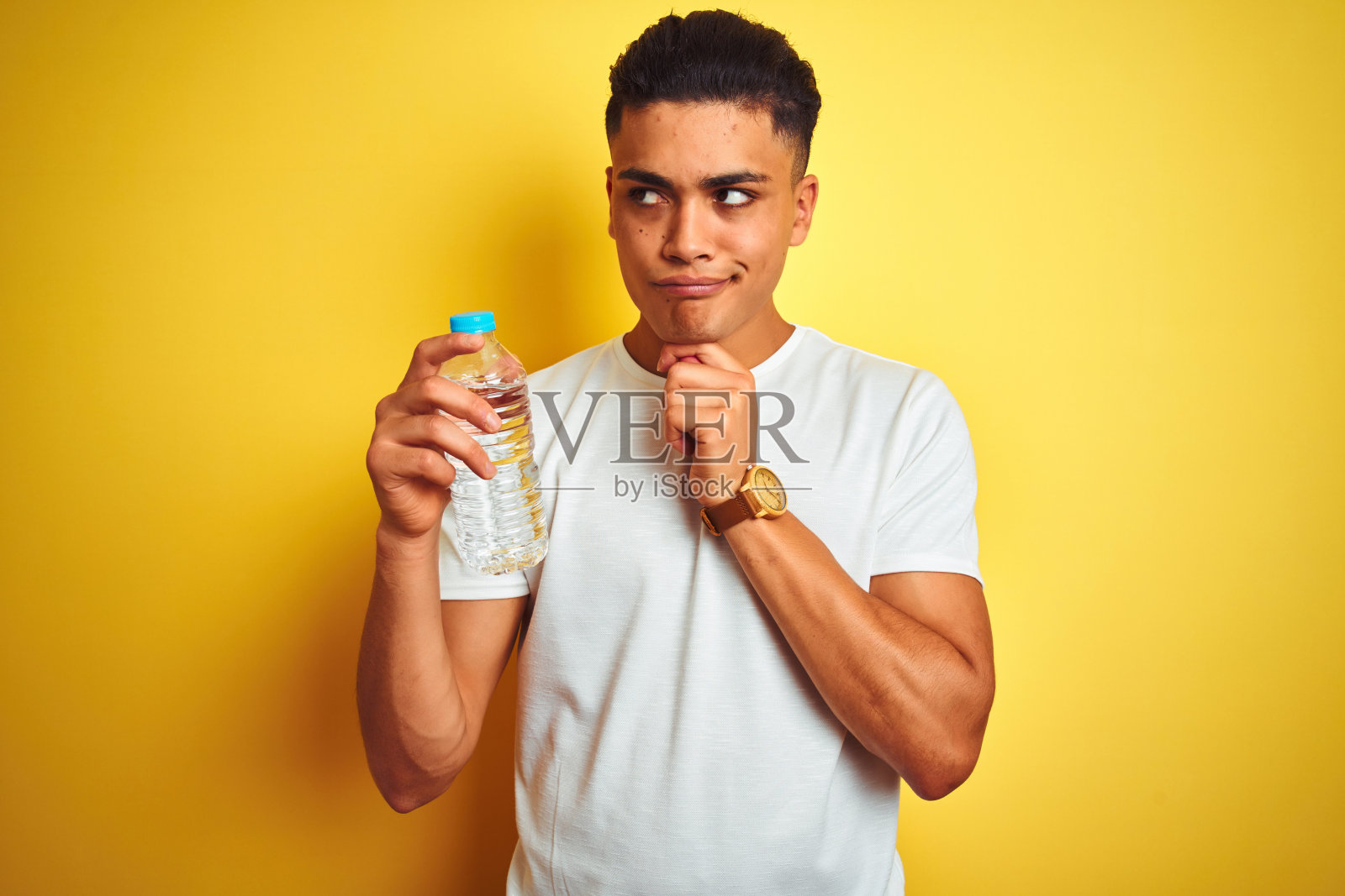 年轻的巴西男子拿着一瓶水站在孤立的黄色背景严肃的脸上思考问题,非常困惑的想法照片摄影图片