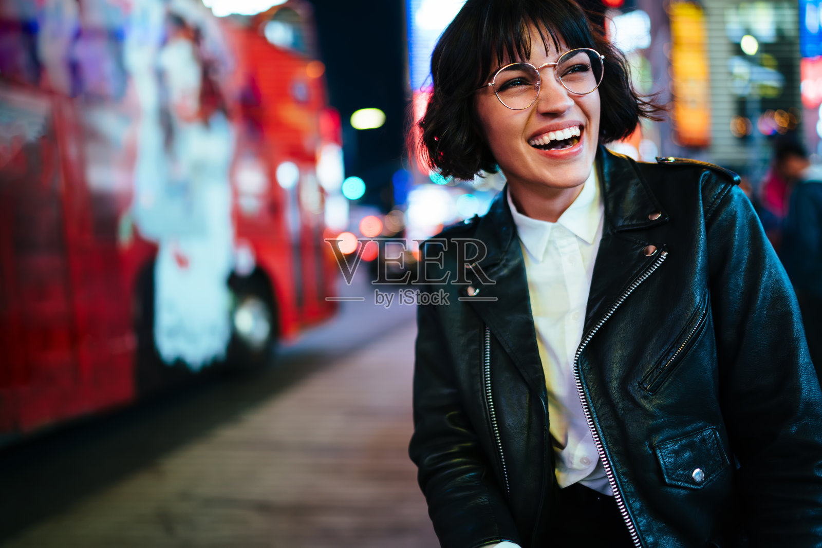 漂亮的积极的年轻女子短发在城市背景下的灯光享受闲暇时间在纽约。快乐的潮人在时尚的穿戴和眼镜有乐趣在大都市的街道照片摄影图片