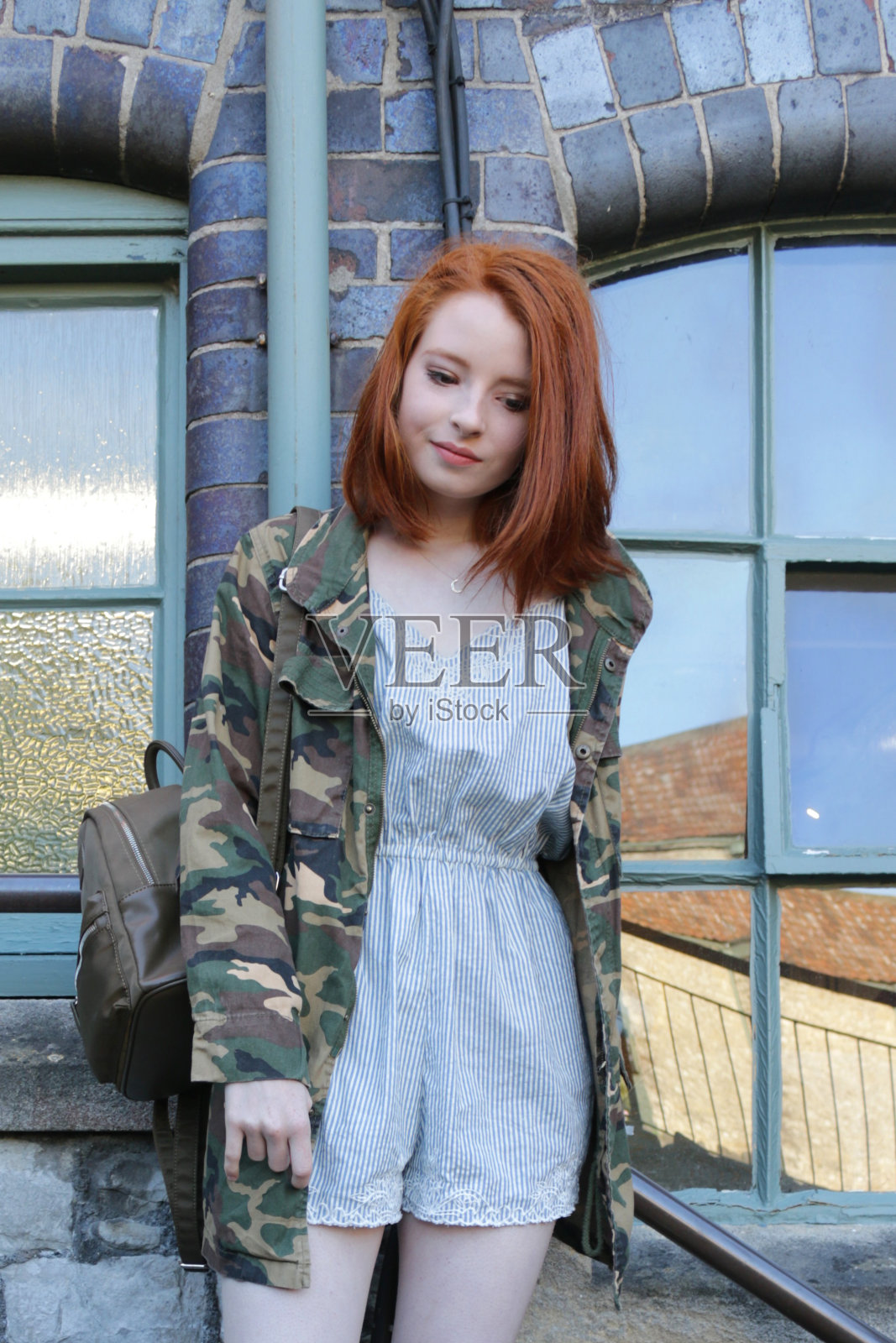 这是一个14 / 15岁的红发少女站在户外工厂的台阶上,穿着一件复古军服外套,外面是一套蓝白条纹的运动服和一双漂亮的米色凉鞋,肩上挎着一个棕色的帆布袋照片摄影图片