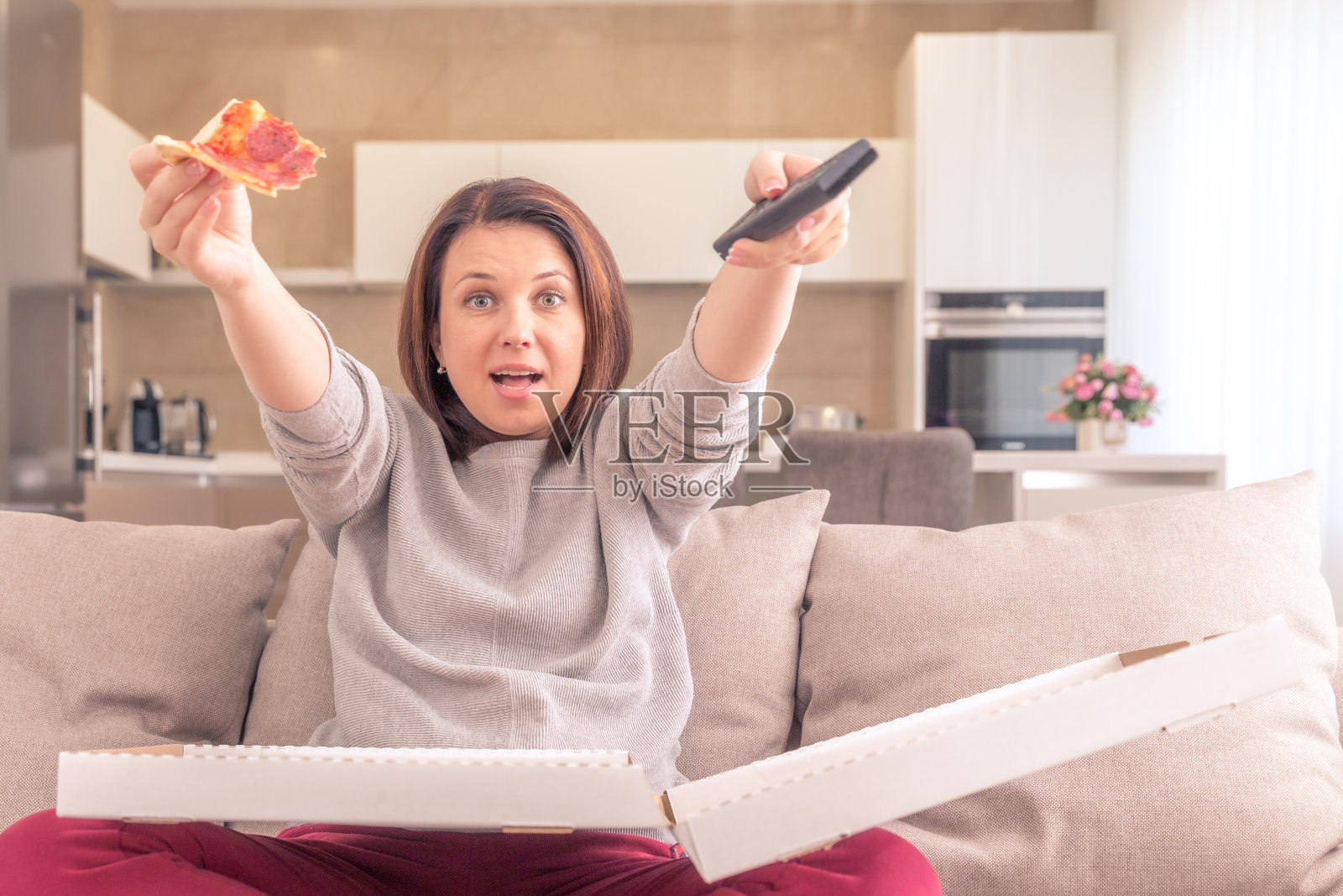 兴奋的女人在家里拿着遥控器边吃披萨边看电视,橙色调照片摄影图片