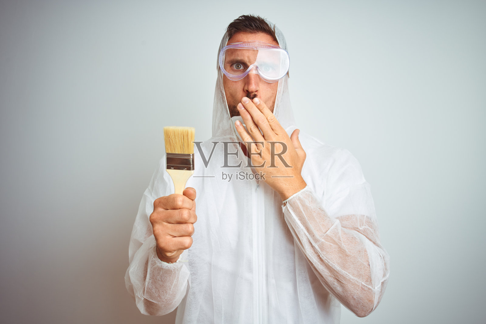 油漆工戴着专业的工人设备,拿着刷子捂在孤立的背景上,用手捂着嘴,因错误而震惊羞愧,表情恐惧,默默害怕,秘密概念照片摄影图片