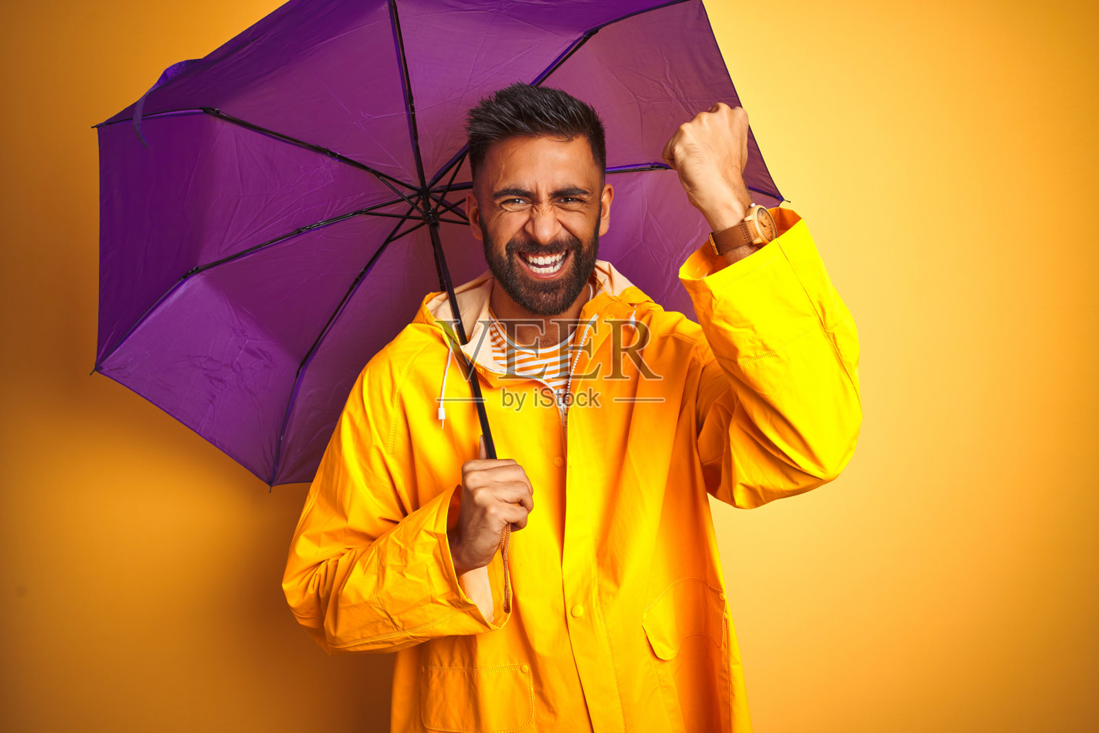 身穿雨衣、打着紫色雨伞的印度青年,在黄色背景前恼怒照片摄影图片