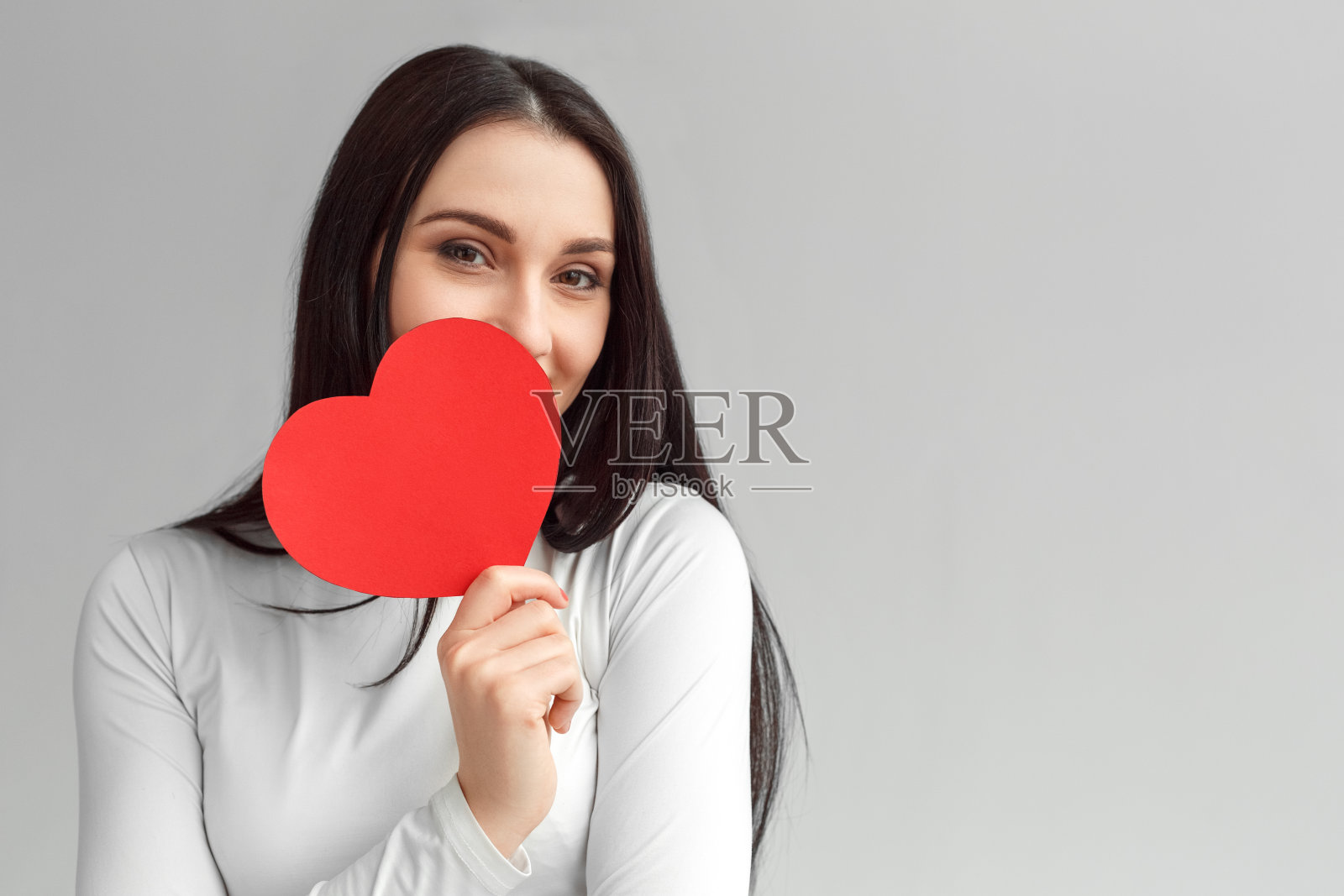情人节。孤立的女子站在灰色的隐藏面孔后面的心卡看着相机好玩的特写照片摄影图片