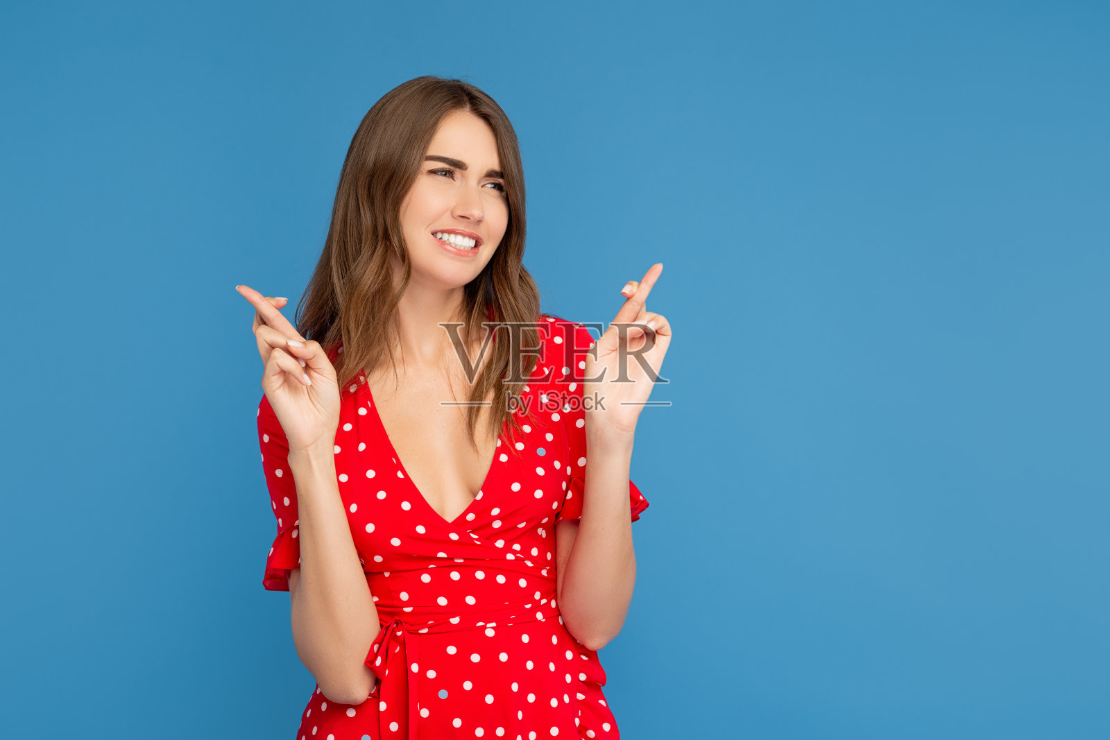 幸福的女人与明亮的微笑在红色的衣服看远离手与手指交叉在希望在蓝色的背景。照片摄影图片