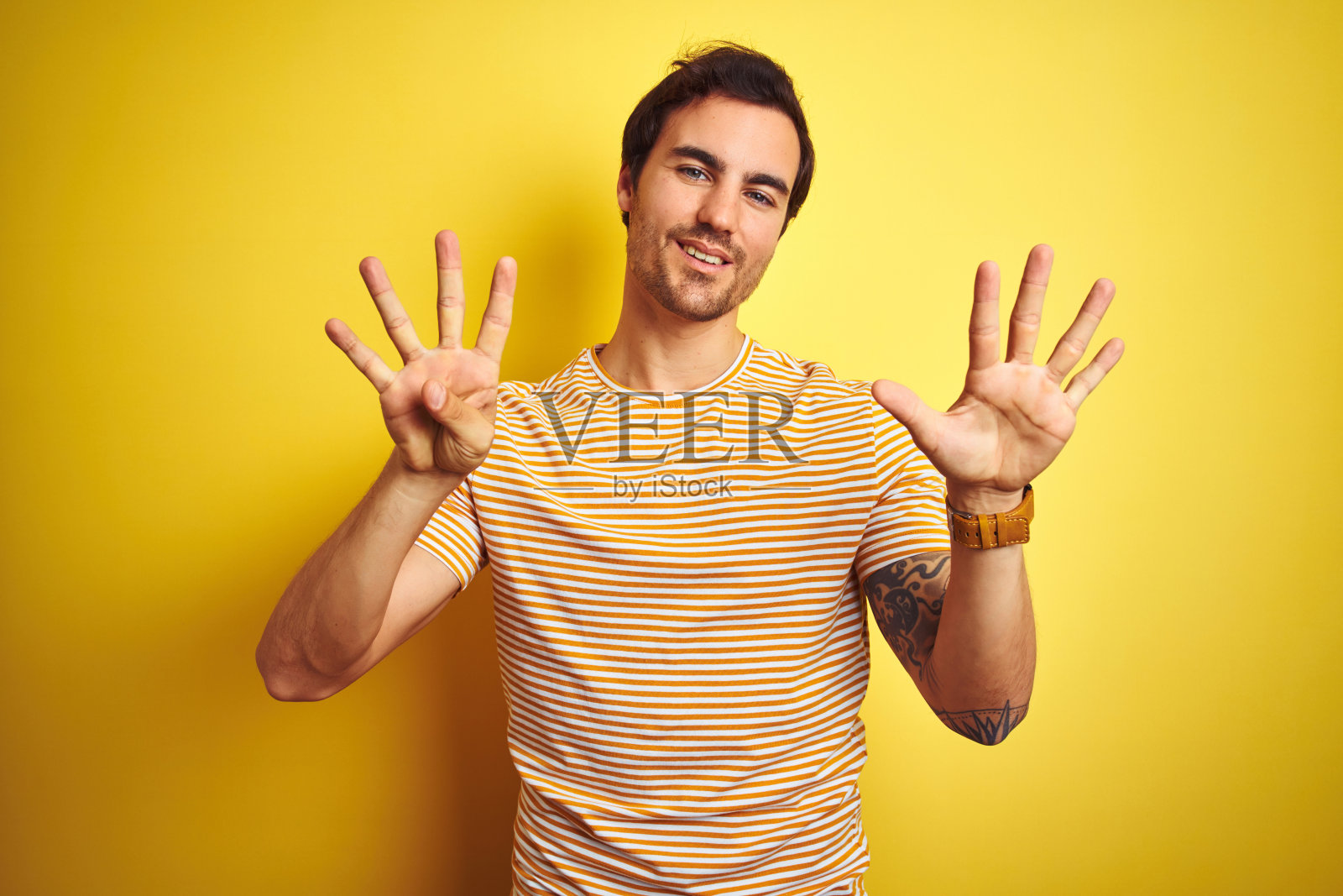 年轻英俊的男子纹身穿条纹t恤在孤立的黄色背景显示和指向数字9,而微笑自信和快乐。照片摄影图片