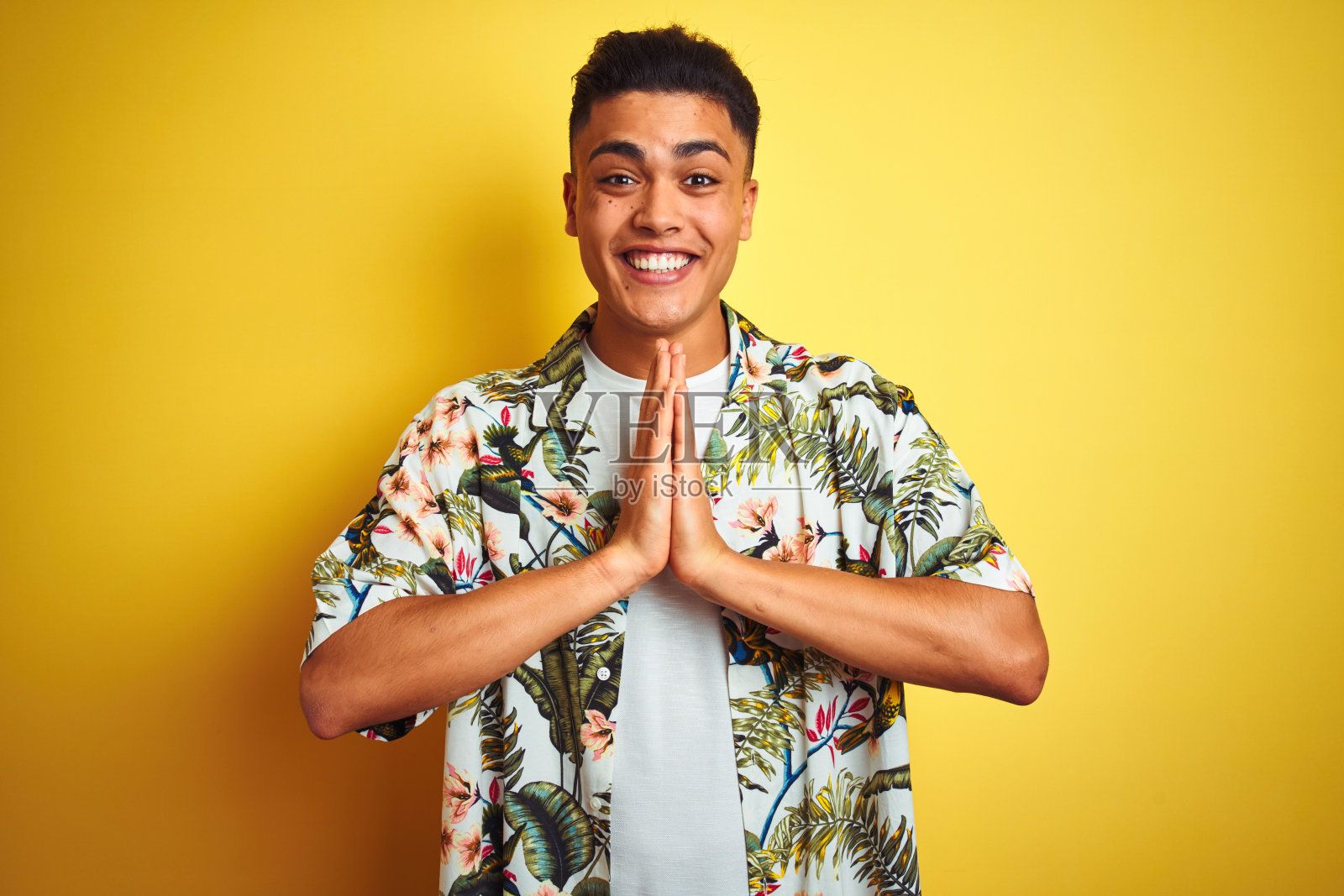 年轻的巴西男子度假穿着夏天的花衬衫在孤立的黄色背景祈祷与手一起请求原谅微笑自信。照片摄影图片