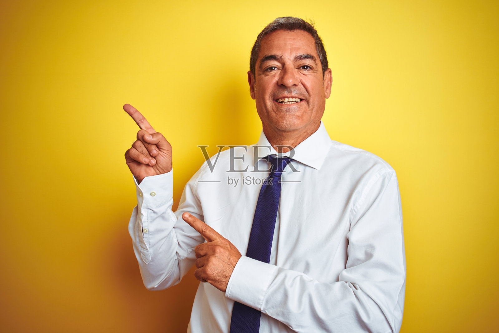 英俊的中年商人站在孤立的黄色背景微笑,看着相机指向与双手和手指的一边。照片摄影图片