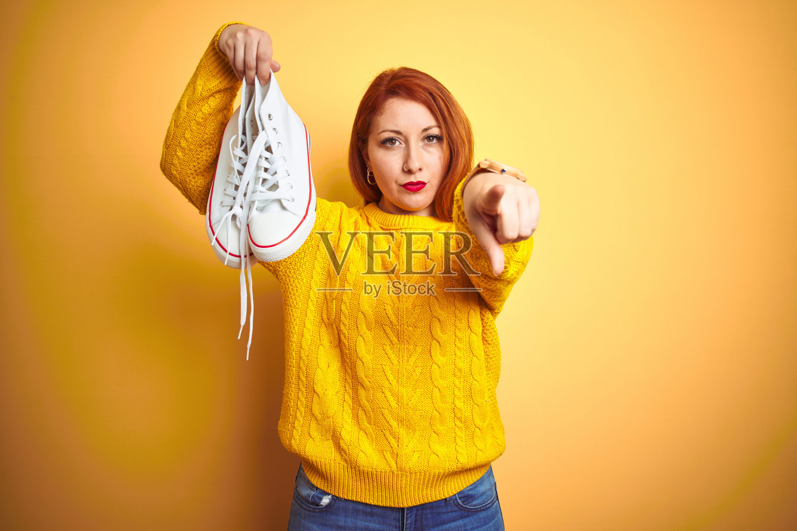 年轻美丽的红发女人拿着白色运动鞋在黄色孤立的背景指向指向相机和你,手势,积极和自信的手势从前面照片摄影图片