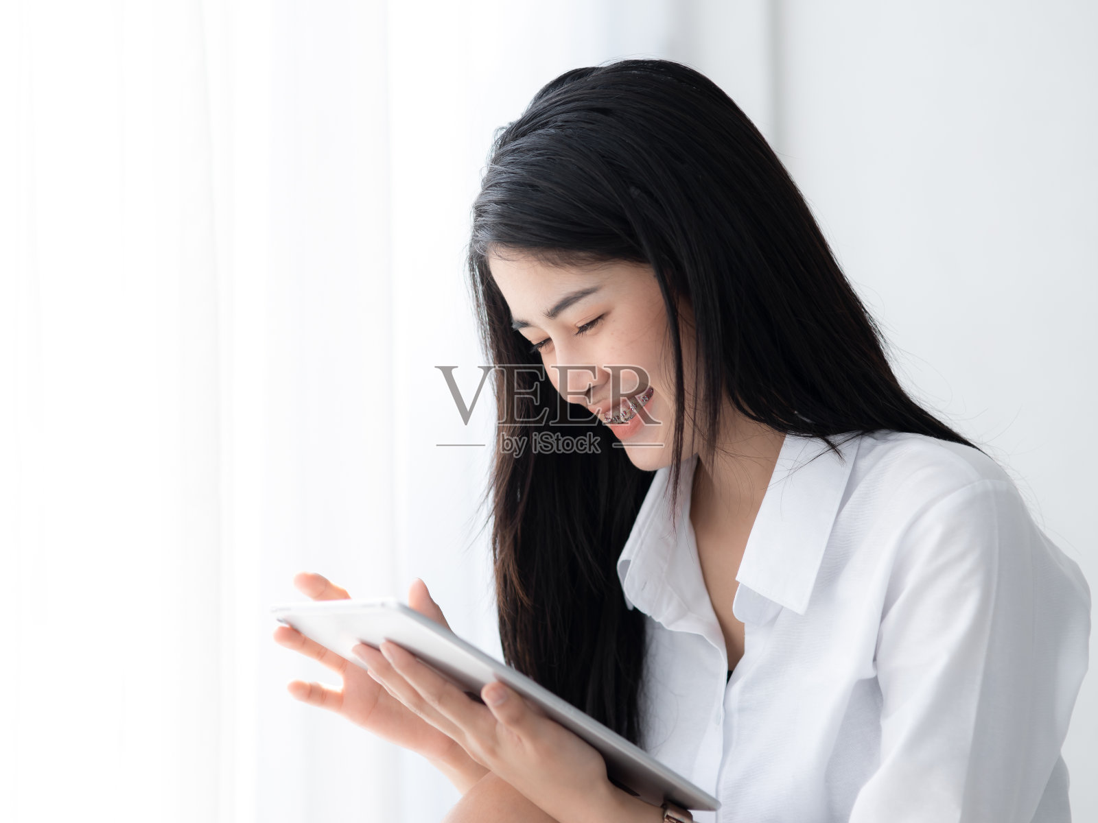 美丽的亚洲女人在白色衬衫使用平板电脑在窗户在早上。照片摄影图片