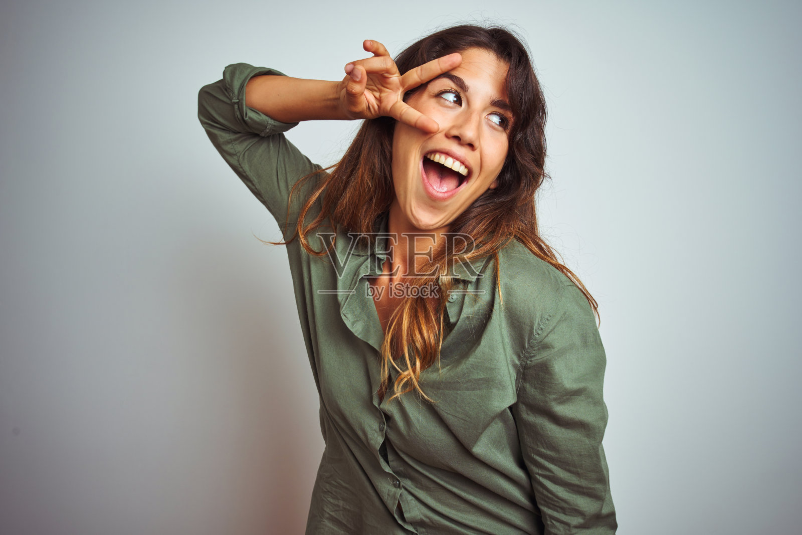 年轻美丽的女子穿着绿色的衬衫站在灰色孤立的背景做和平的象征,手指在脸上,微笑着欢快的胜利照片摄影图片
